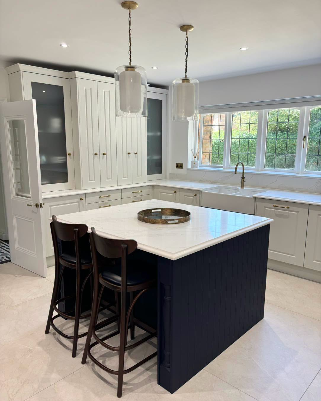 Modern kitchen with white cabinets, navy island, pendant lights, and three bar stools by a large window.