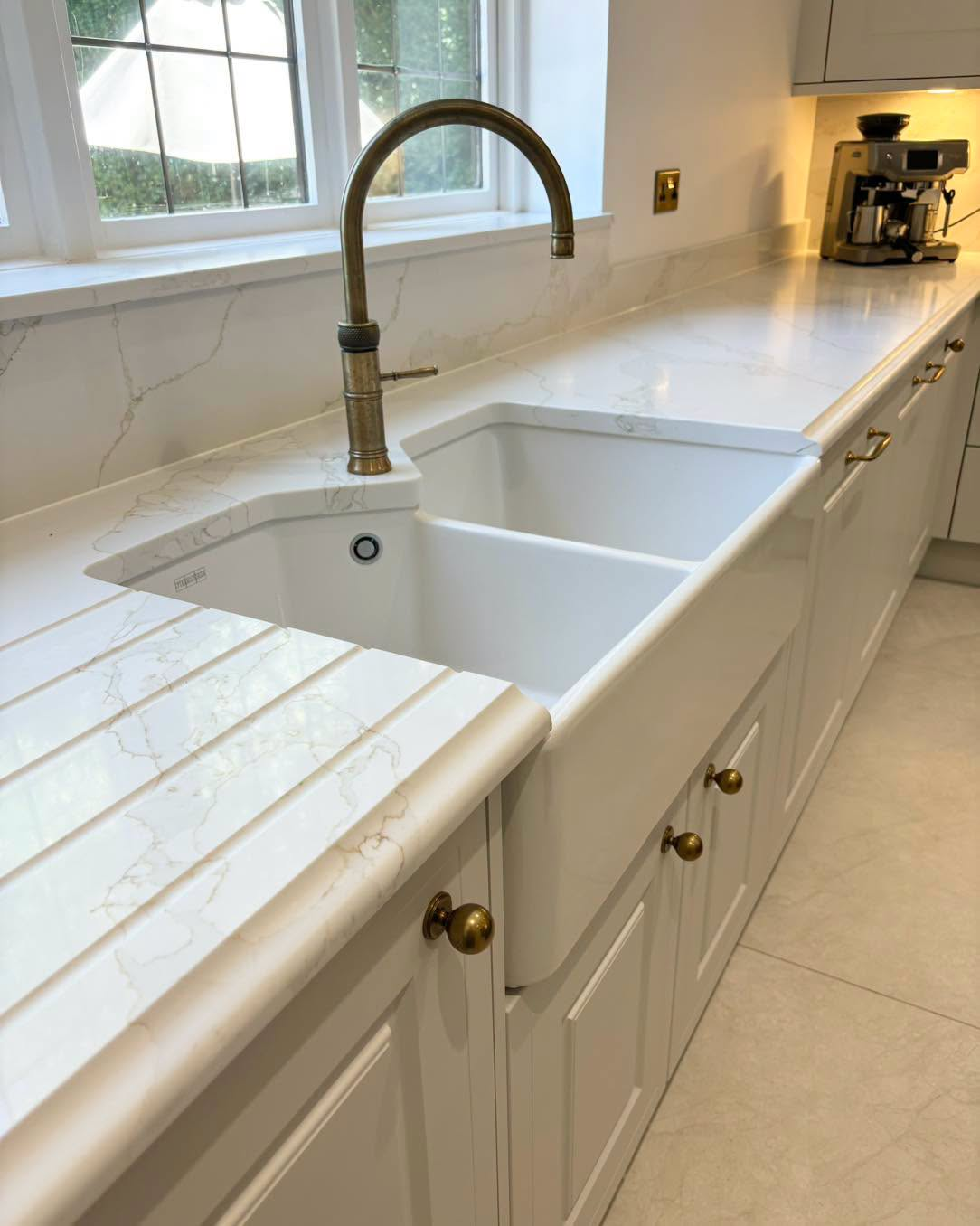 White double farmhouse sink with brushed brass faucet in a bright kitchen with marble countertop