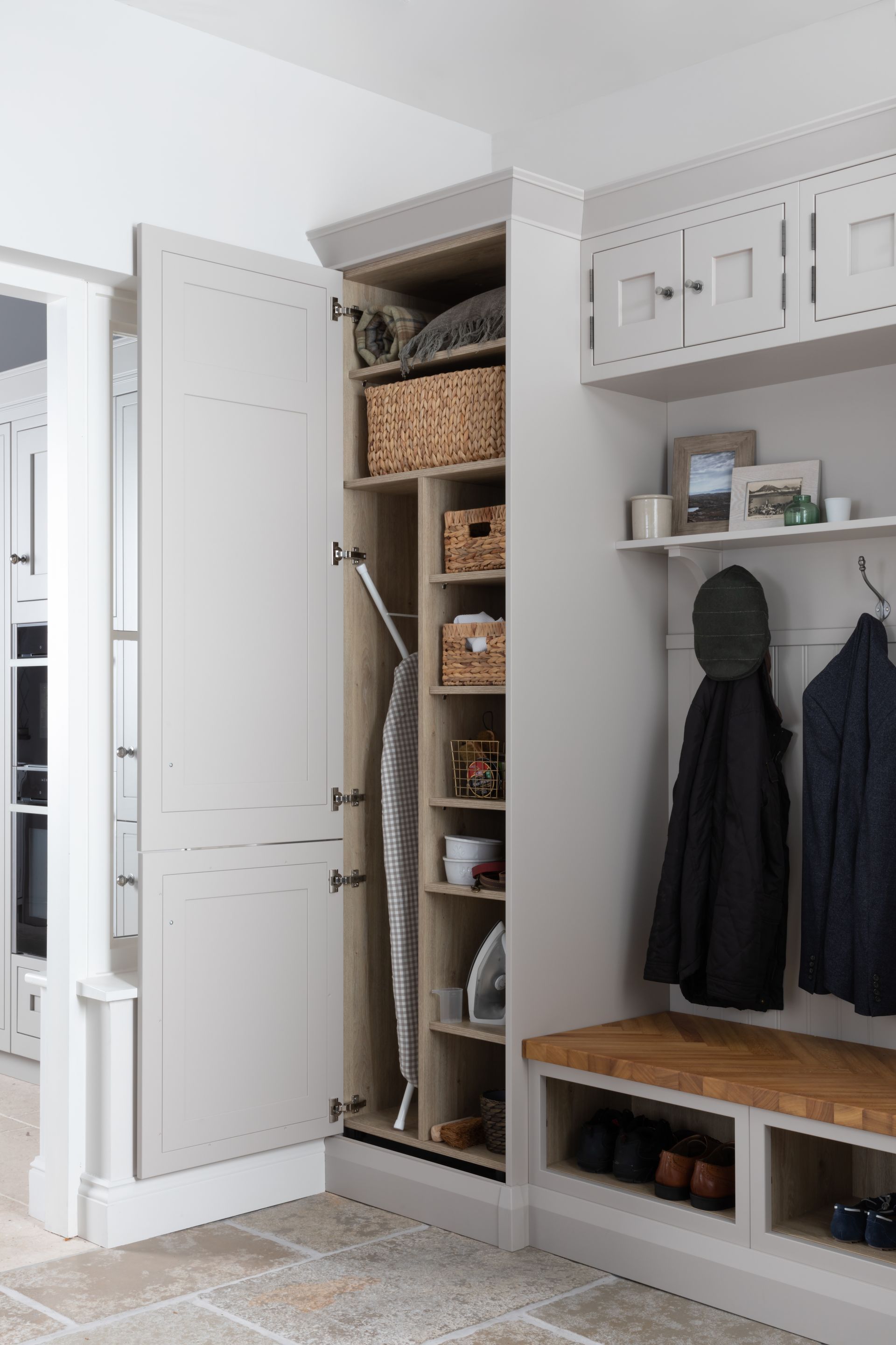 A light gray, built-in mudroom storage unit with a tall cabinet door open, revealing an ironing board and tiered shelves.