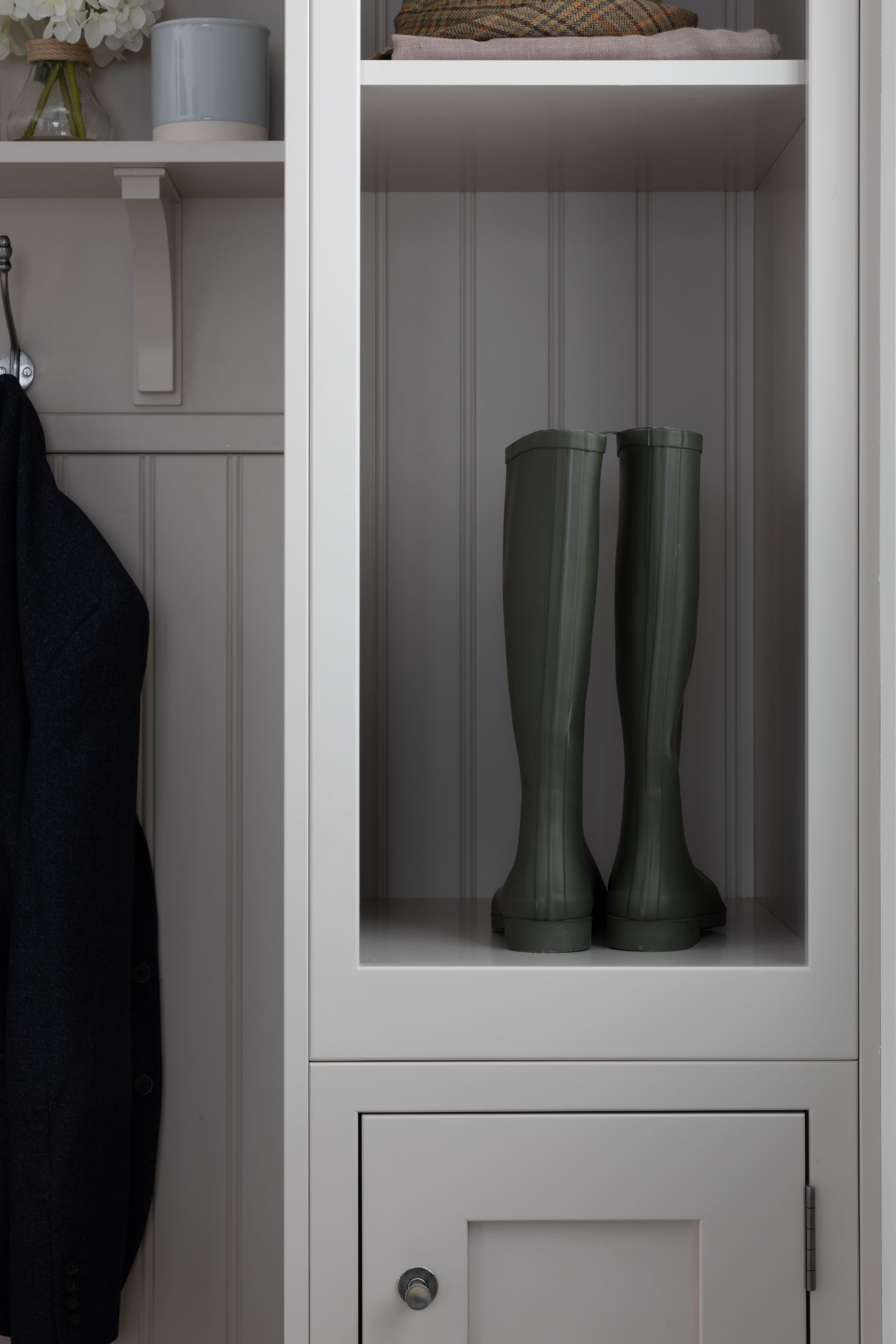 A pair of olive green rubber boots sit on a shelf in a white, built-in mudroom storage unit next to a hanging coat.