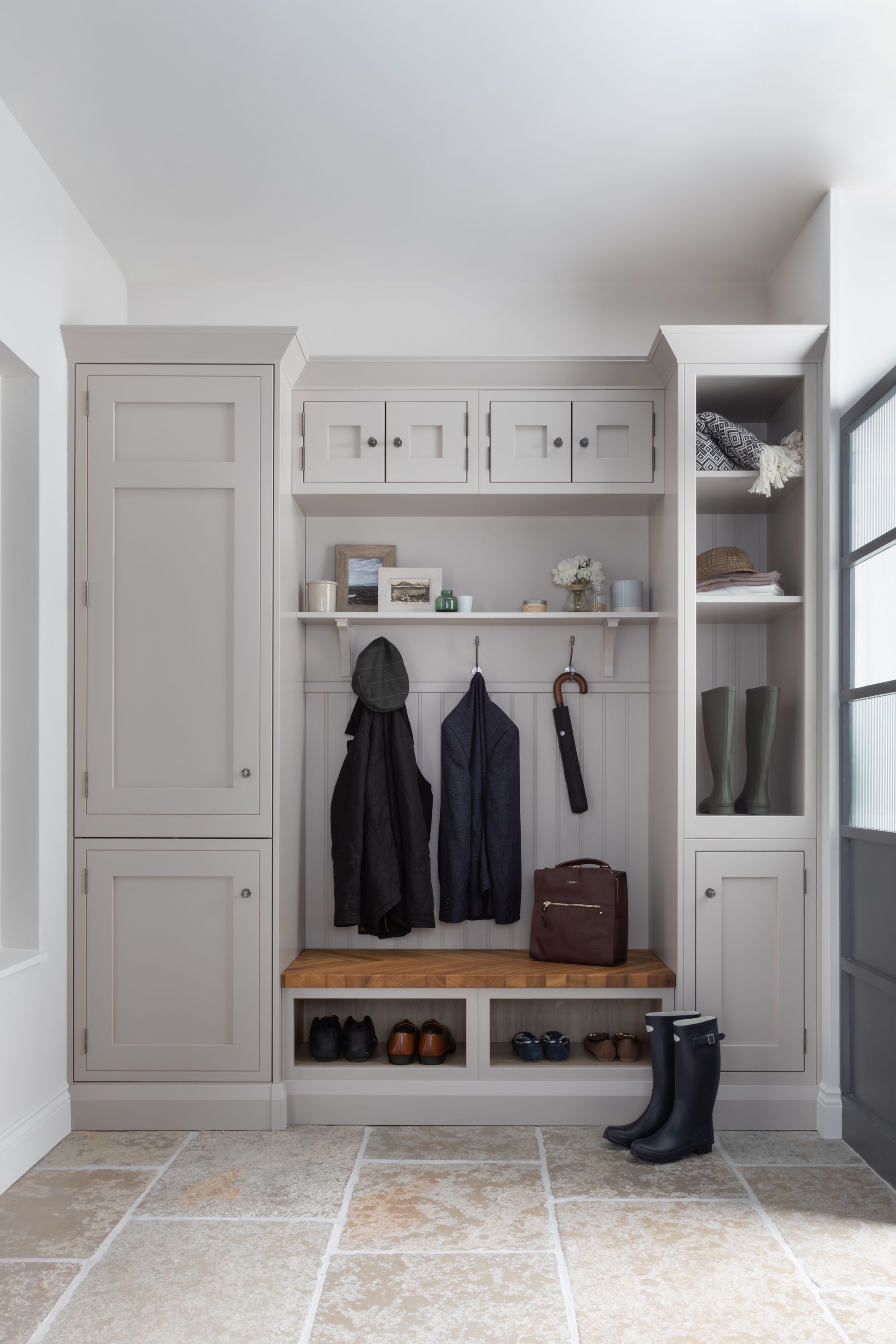 A light gray built-in mudroom storage unit with a wooden bench, coat hooks, cabinets, shelves, and stone tile flooring.