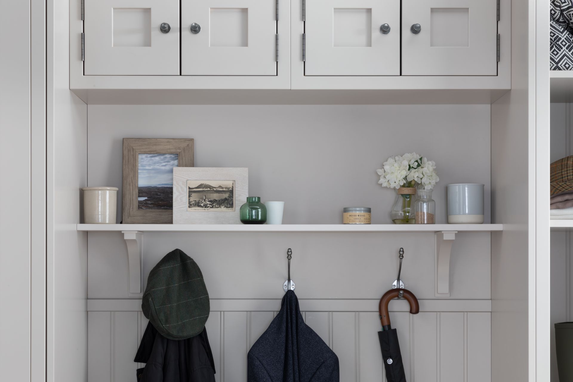 A white entryway mudroom unit with upper cabinets, a shelf holding decor, and coat hooks featuring hats and an umbrella.