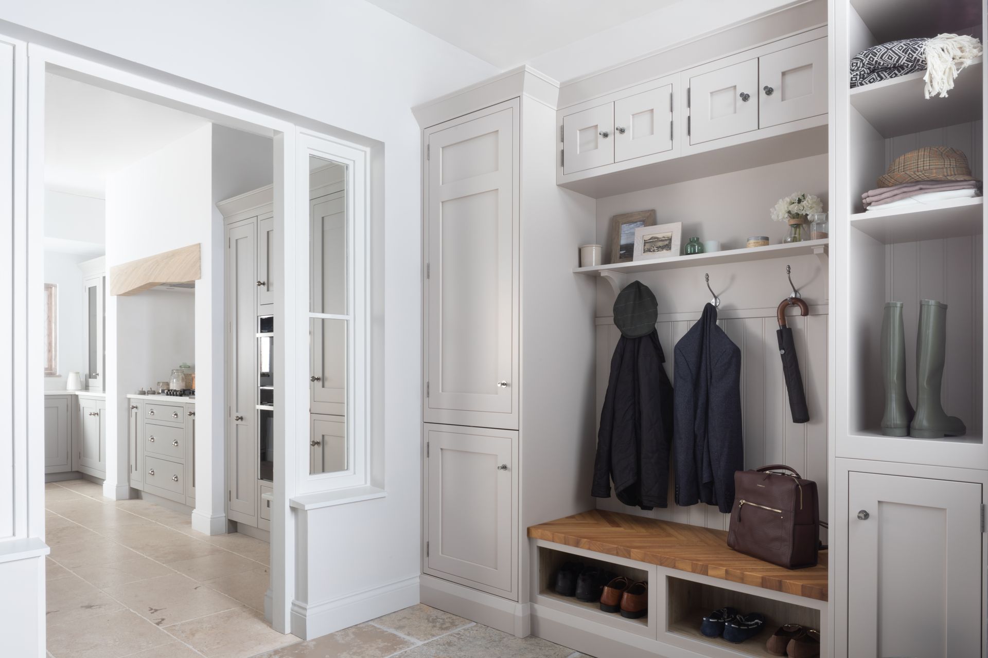 A light-gray entryway mudroom featuring built-in cabinetry, coat hooks, a wooden bench, open shelving, and boot storage.