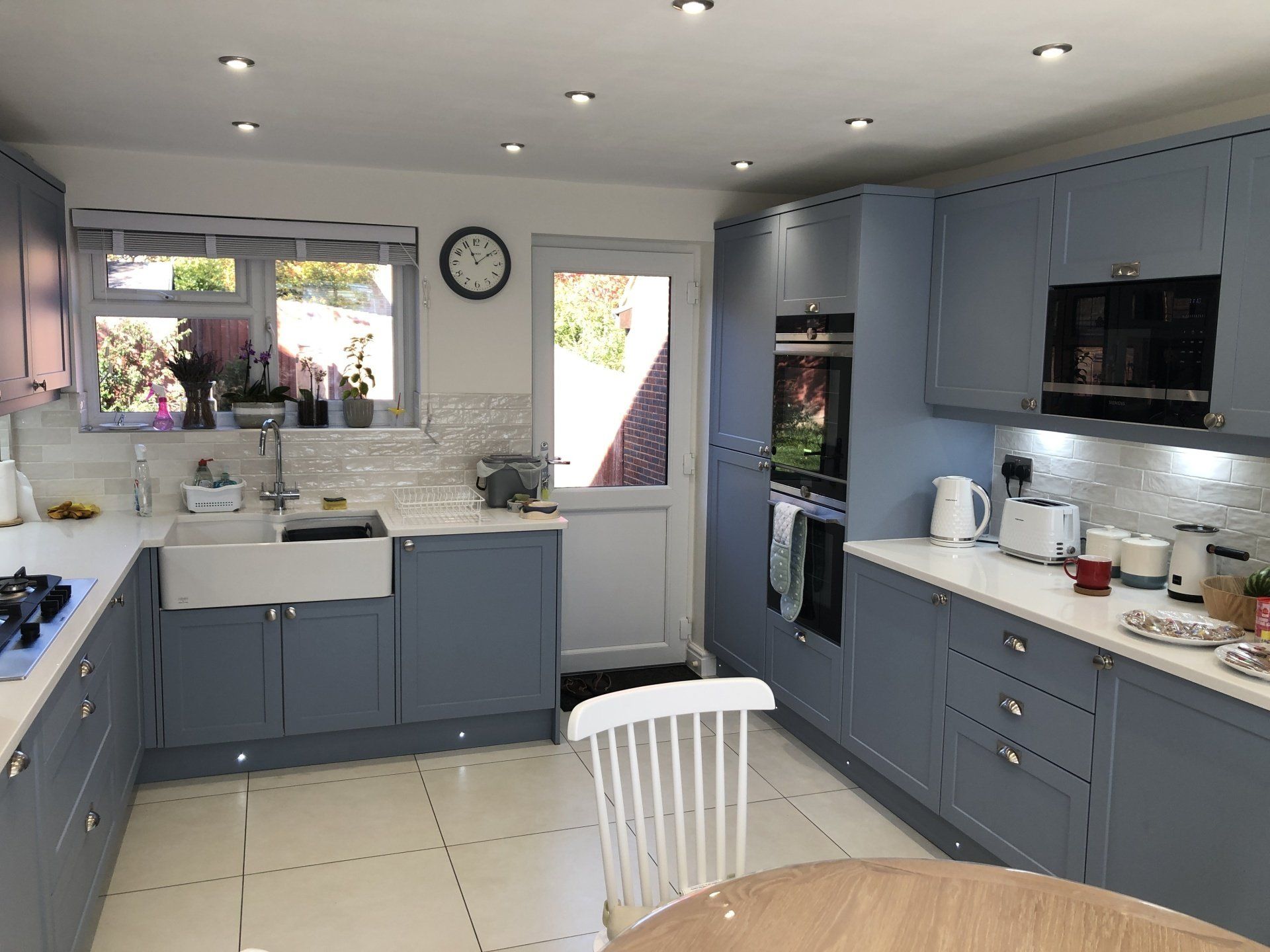 Modern kitchen with blue cabinets, white countertops, a farmhouse sink, and a view of a garden through a door and window.