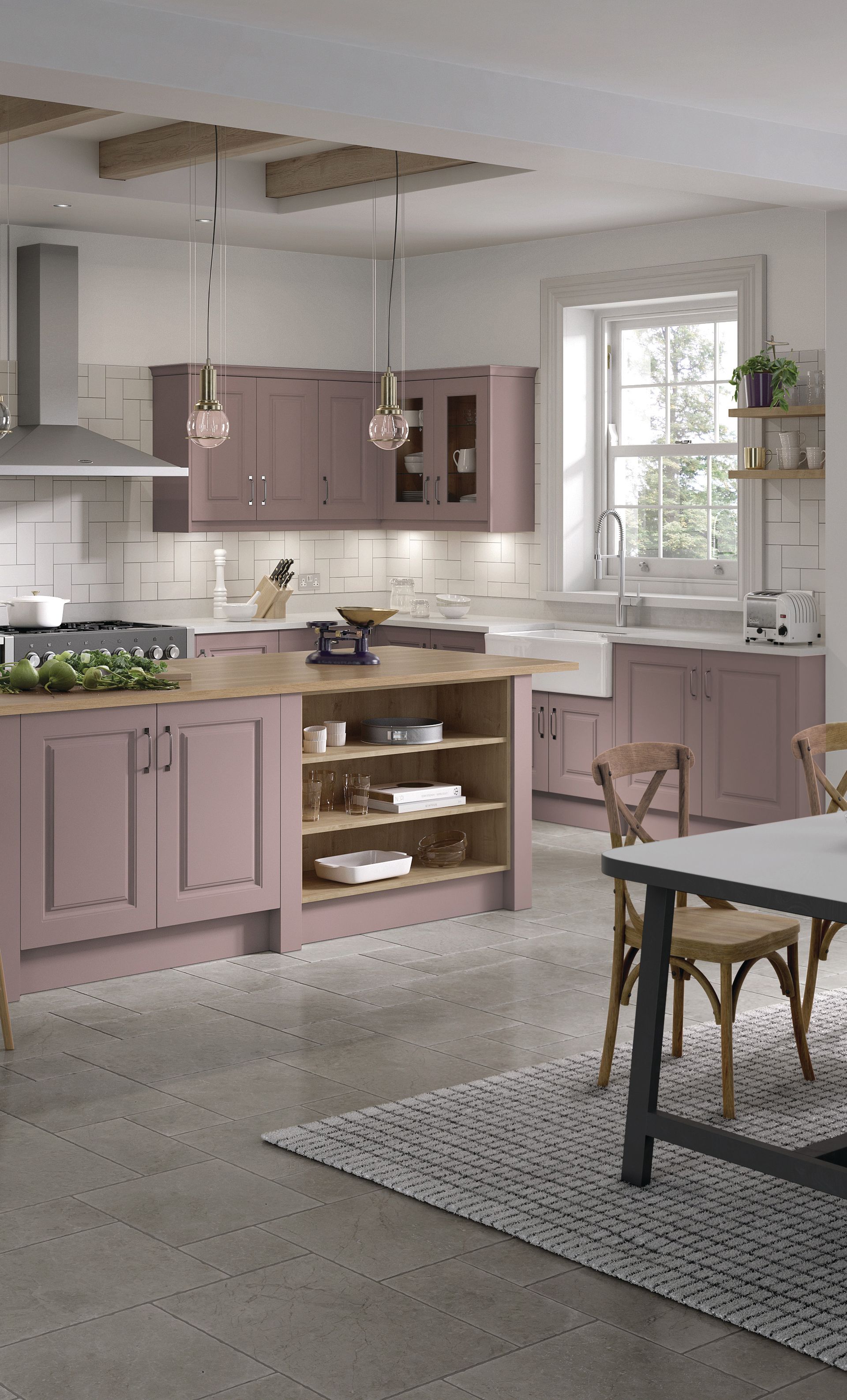 A kitchen with mauve cabinets, a wood-topped island, light stone floors, and a dining area with a patterned rug.