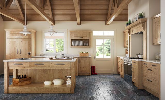 A rustic kitchen with light wood cabinets, an island, vaulted timber ceilings, and dark stone flooring.