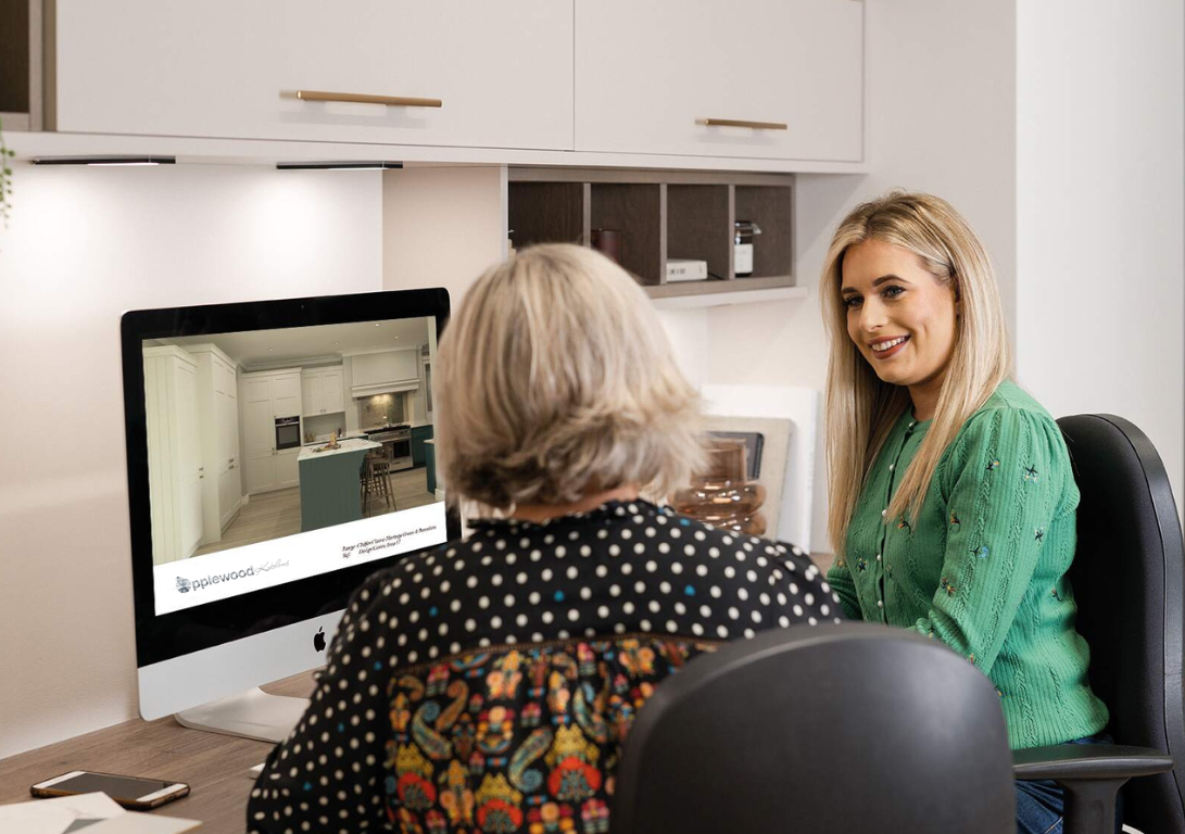 Two people sit at a desk, looking at a computer monitor displaying a 3D design of a modern kitchen interior.
