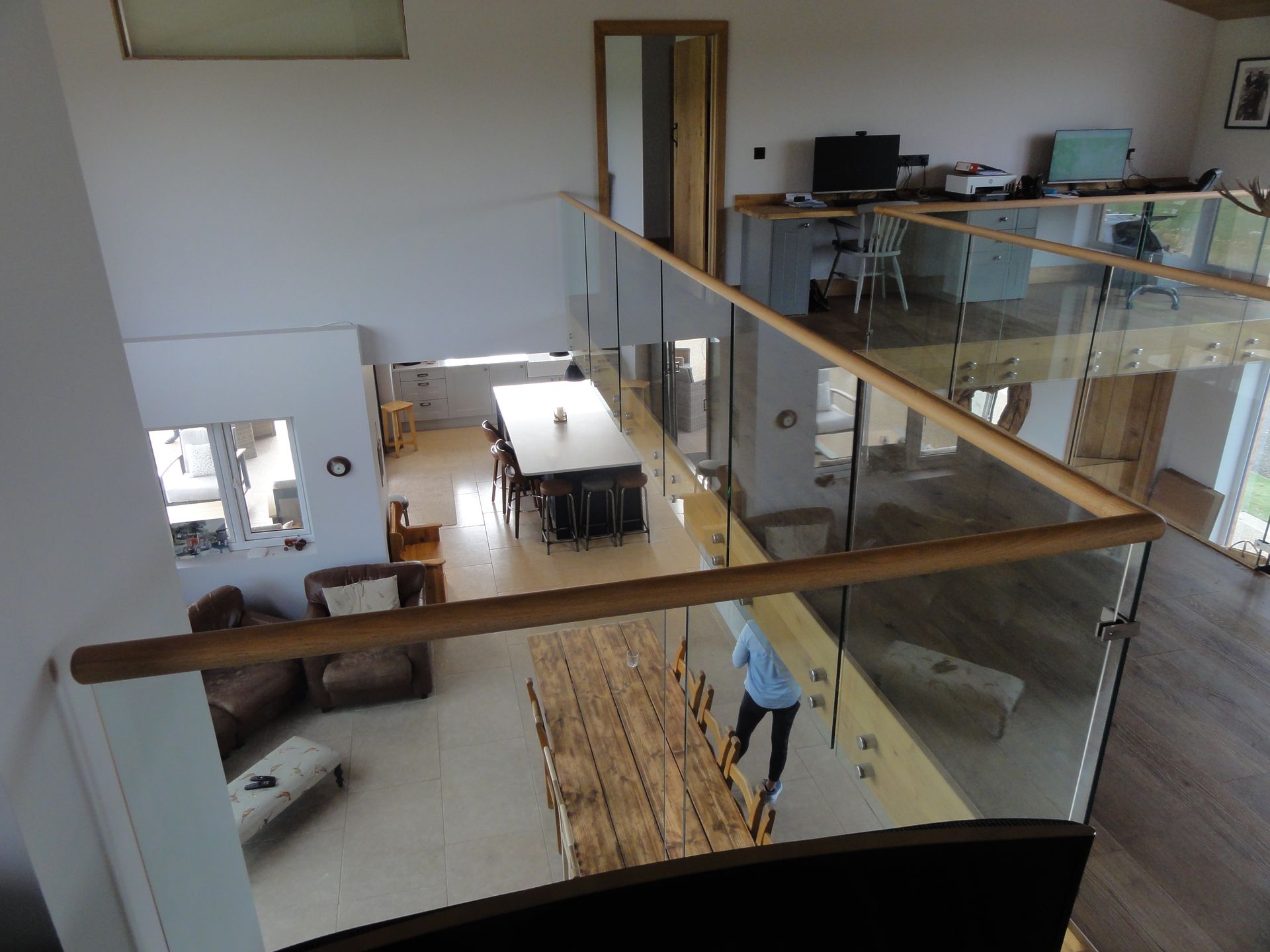 A high-angle view looking down from an indoor glass balcony onto a living area with a long dining table and chairs.
