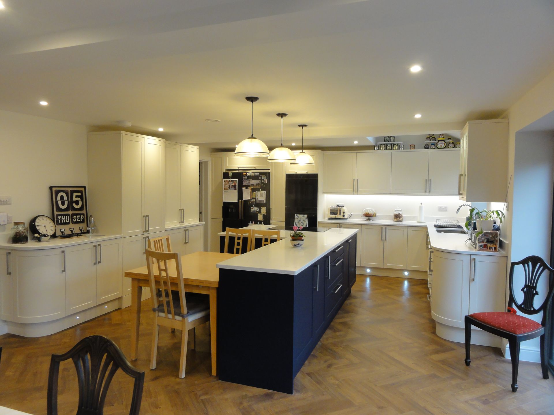 A modern kitchen featuring a dark blue island with a wooden table, white cabinetry, and herringbone wood flooring.