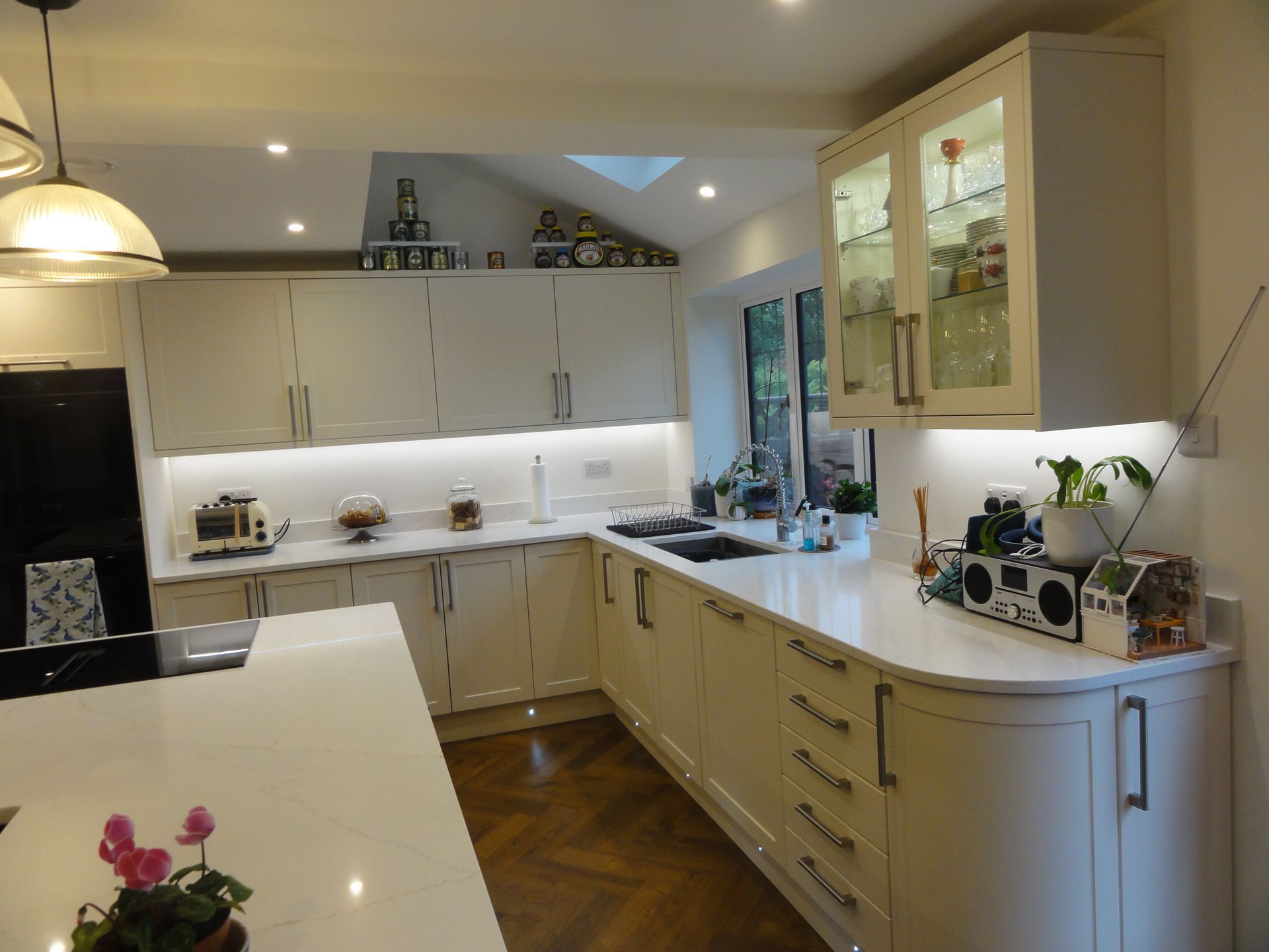 A bright modern kitchen with cream cabinets, white countertops, a central island, and herringbone wood flooring.