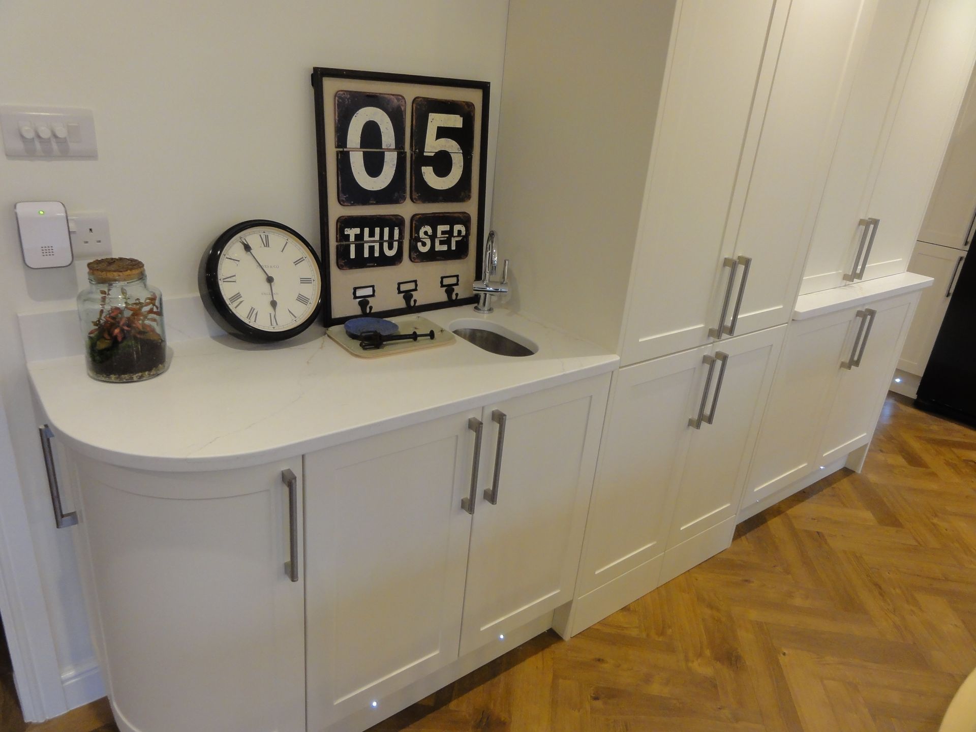 A modern kitchen unit with white cabinetry, a countertop, a decorative wall calendar, a clock, and a glass jar.