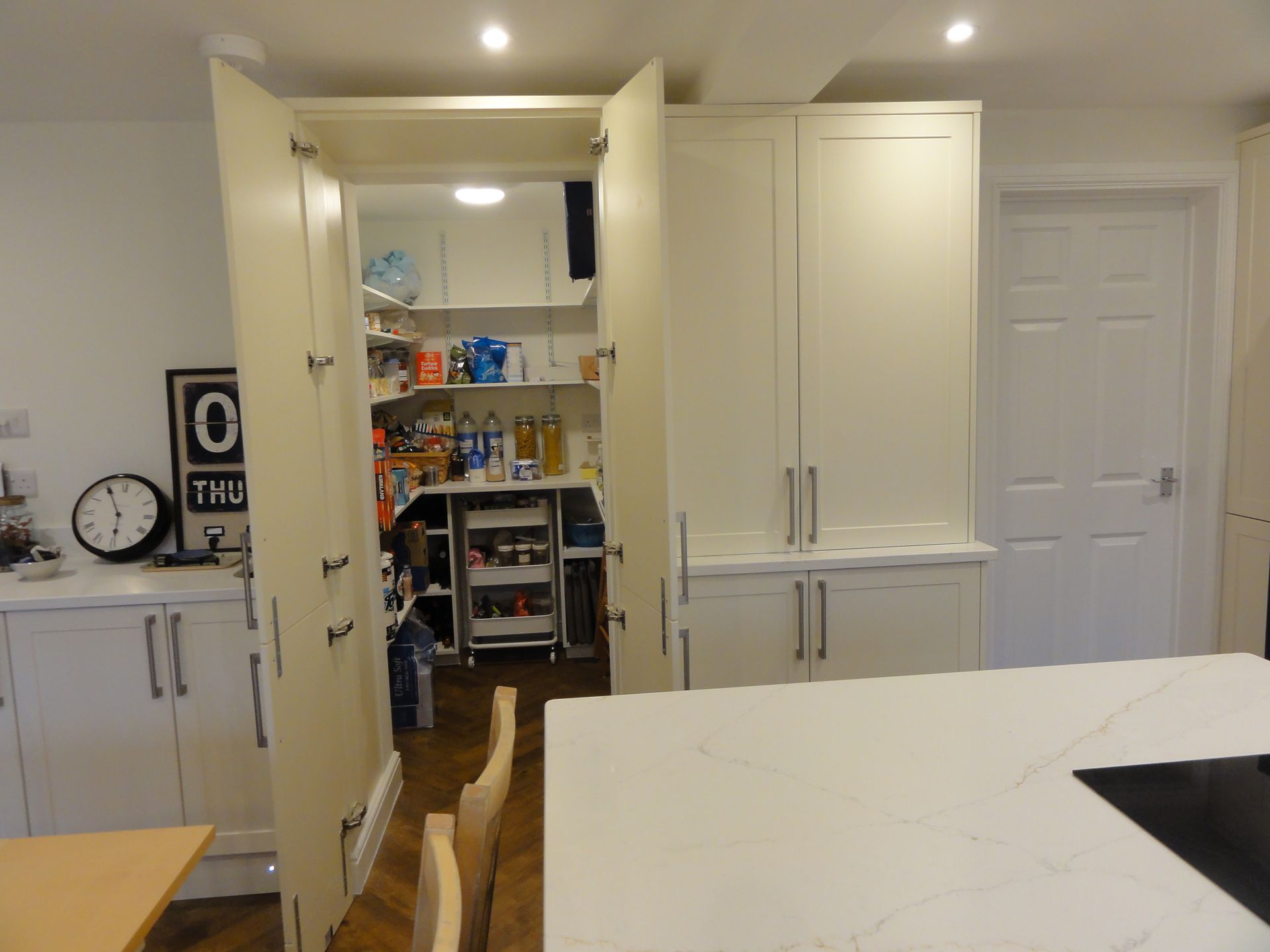 Open double doors reveal a organized walk-in pantry with shelving and items inside a bright, modern kitchen.