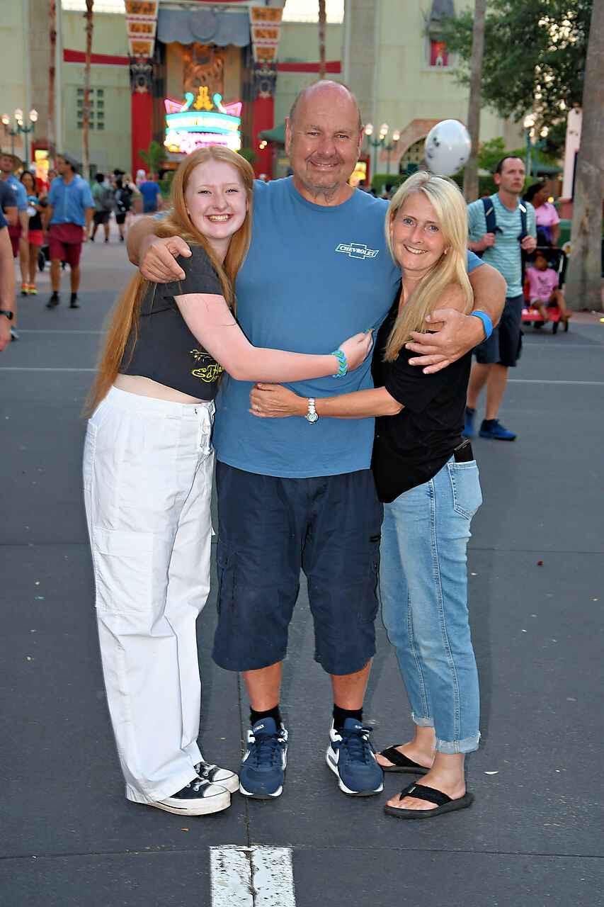 Three people hug in a theme park, smiling for a photo in front of a building entrance on a sunny day.