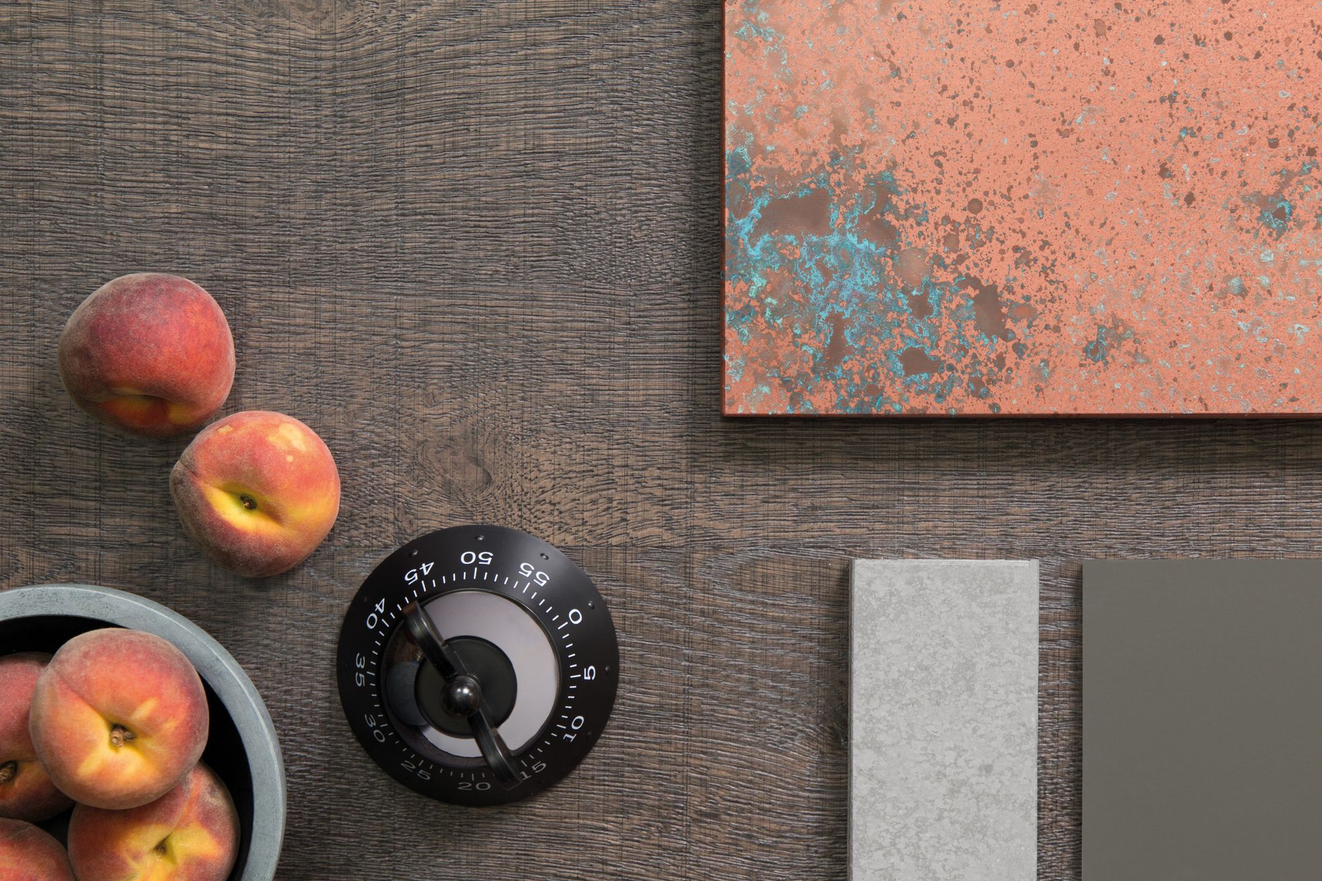 A flat lay features a black kitchen timer, three peaches, a copper-patina panel, and two grey sample tiles on dark wood.