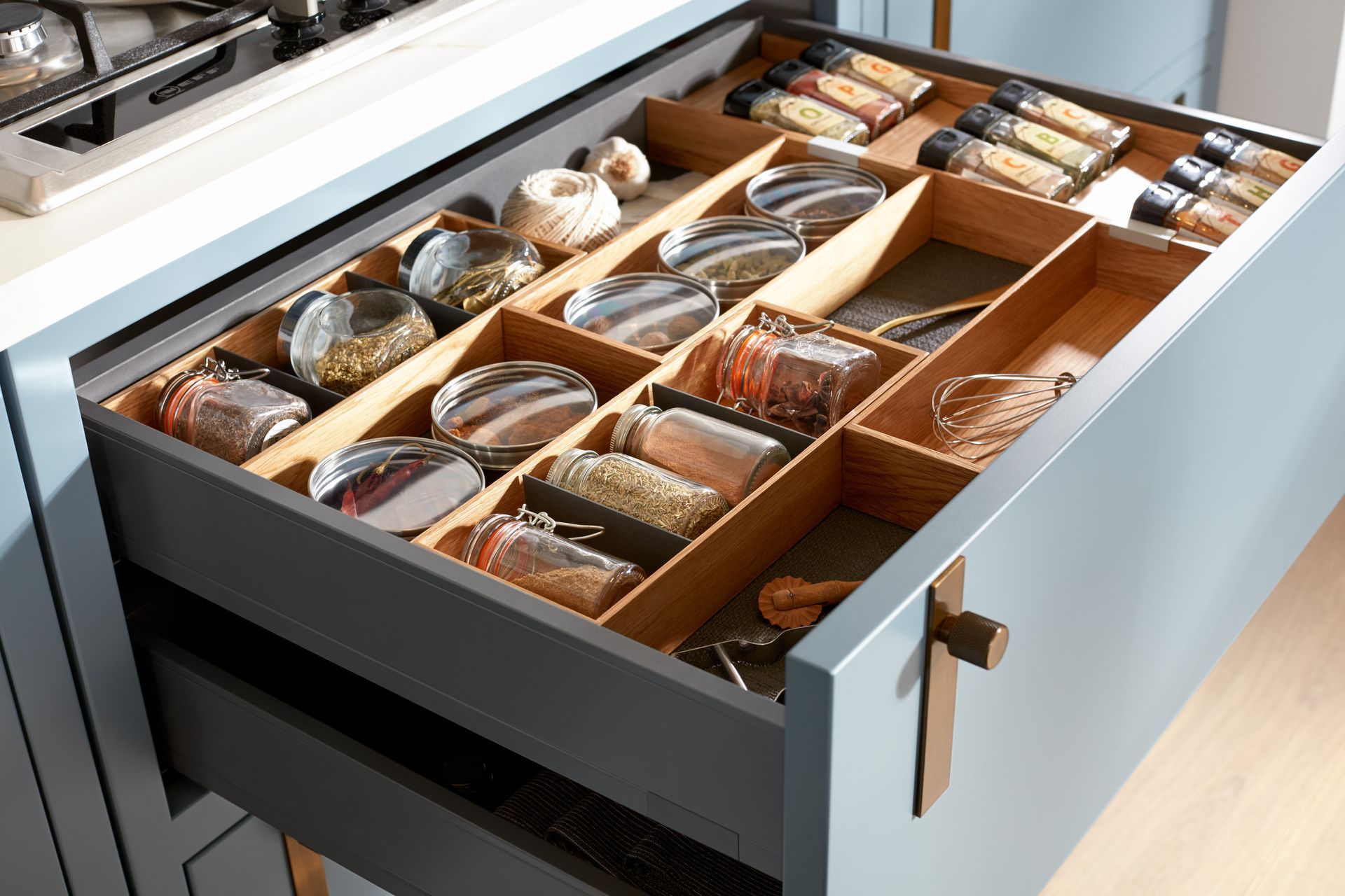 An open blue kitchen drawer with light wood dividers, organized with glass jars, spices, and cooking supplies.