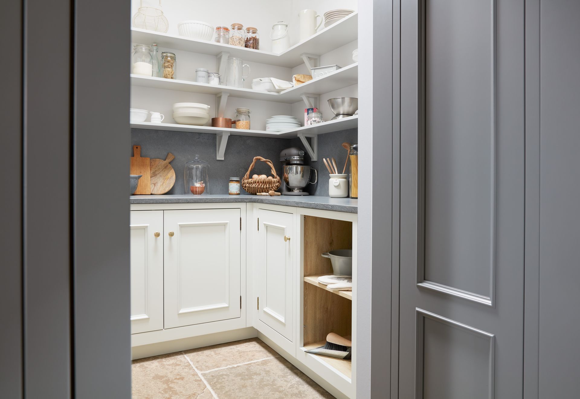 A butler's pantry featuring white cabinets, grey countertops, corner shelves with kitchen items, and light stone flooring.