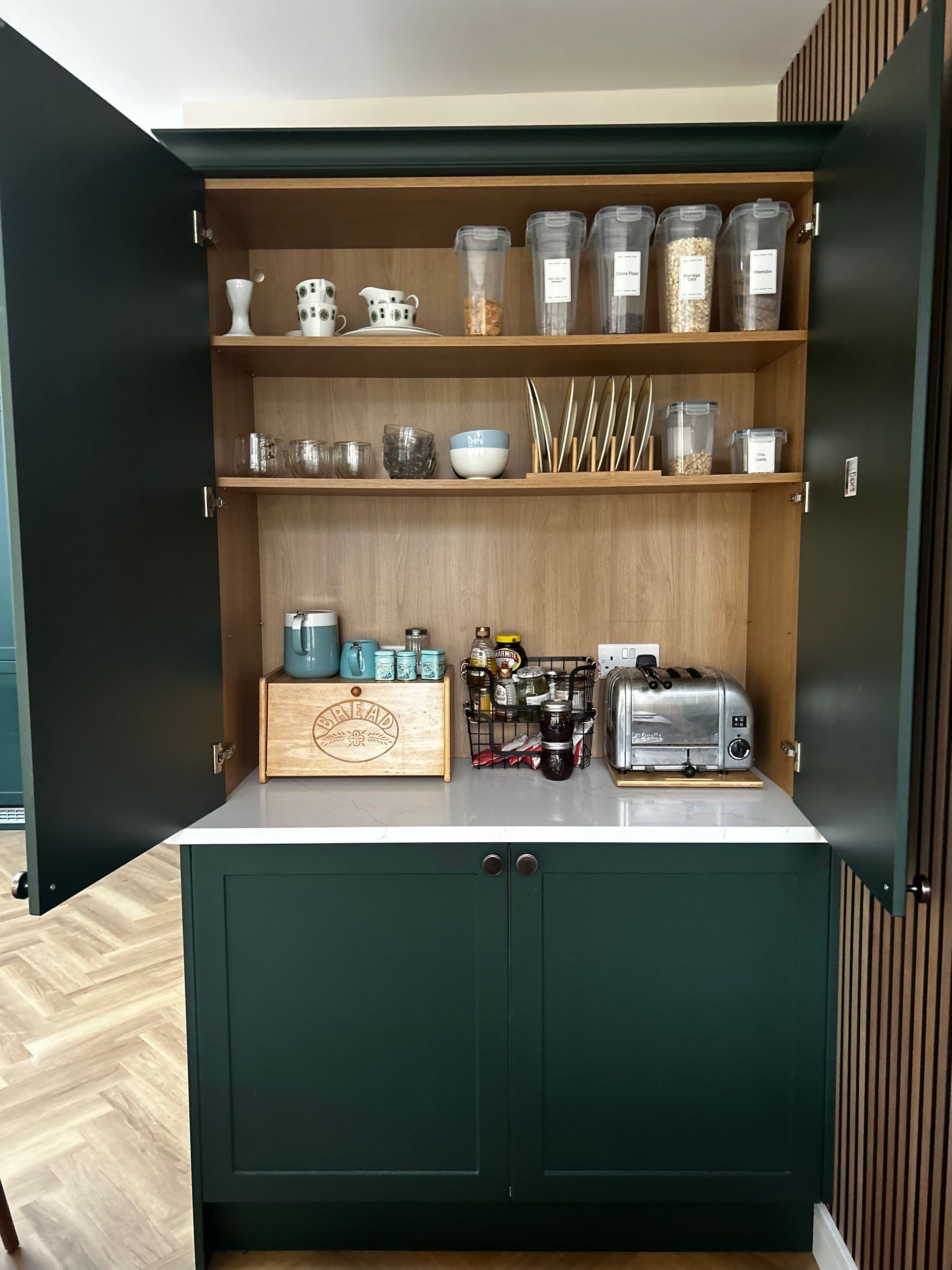 A dark green, open pantry cabinet with wooden interior shelving filled with food containers, kitchen items, and a toaster.