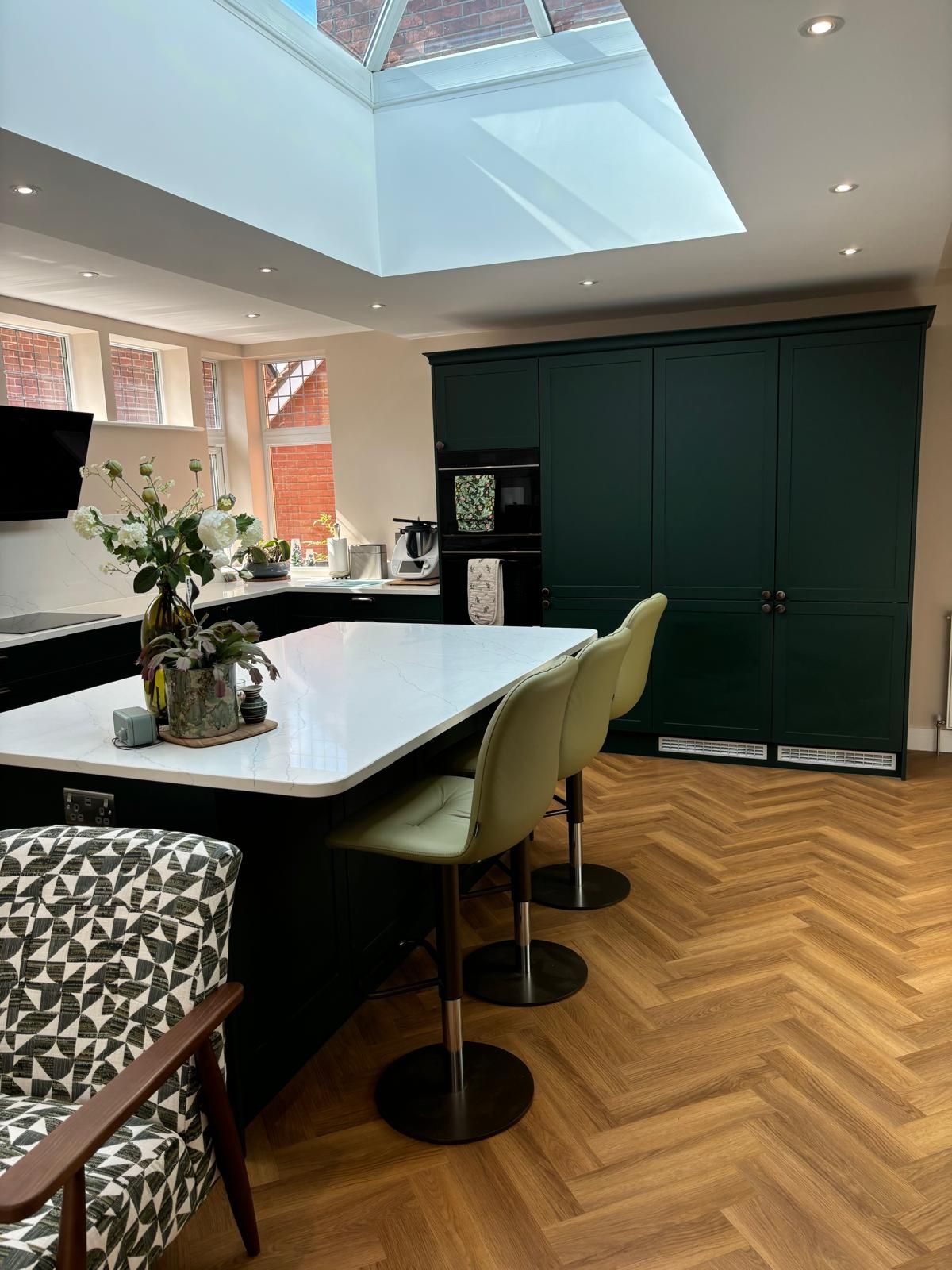 A bright kitchen with a white island, olive green bar stools, dark green floor-to-ceiling cabinets, and parquet flooring.