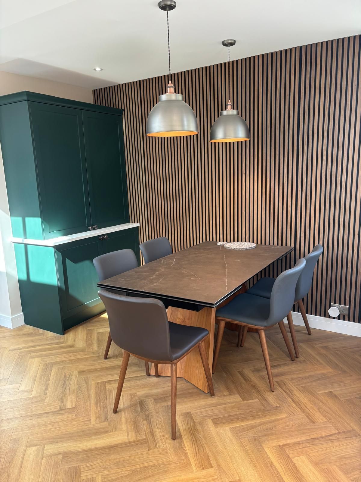 A dining area with a wood-slat wall, a dark wooden table, grey chairs, two pendant lights, and a dark green cupboard.