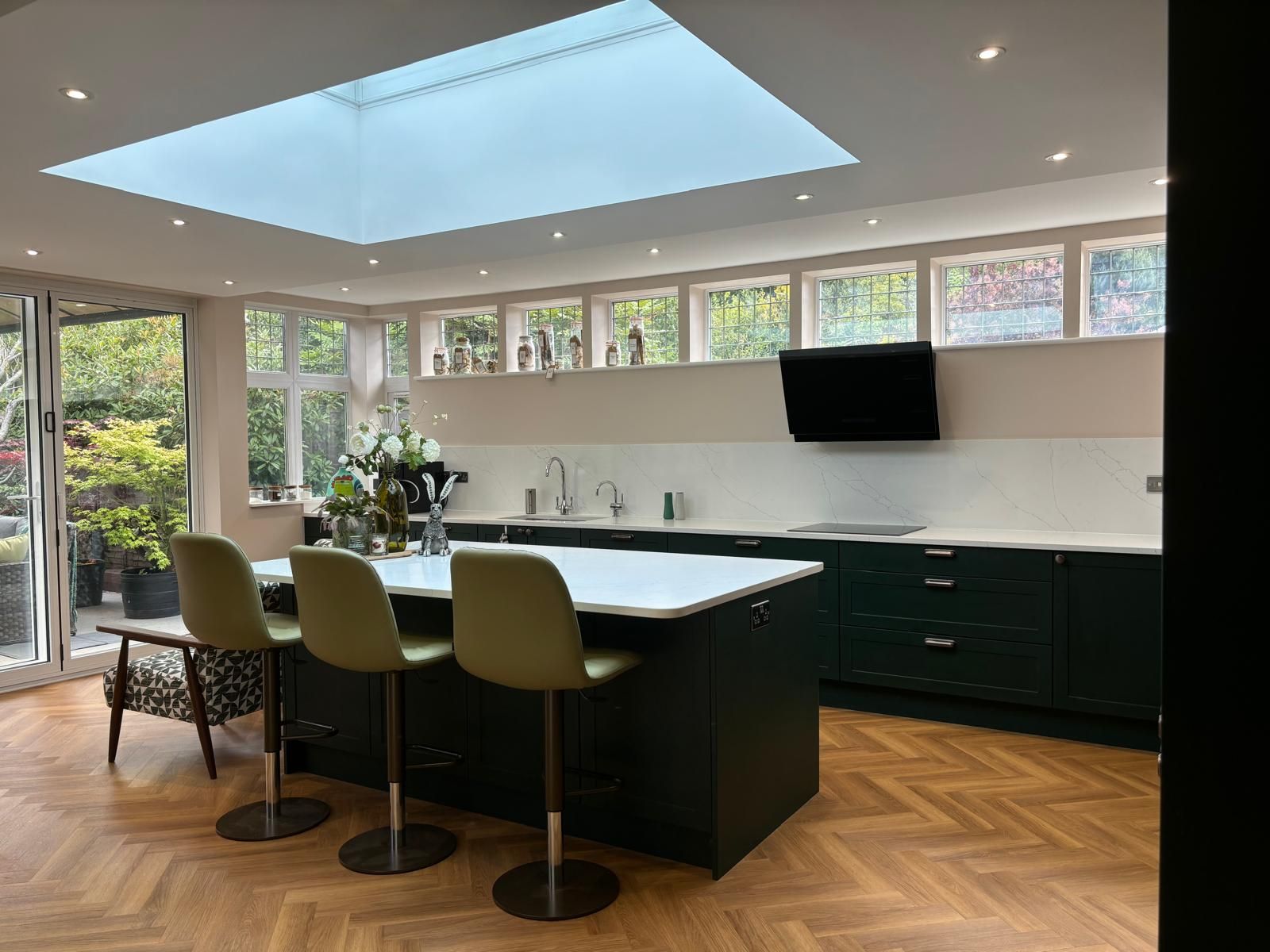 A kitchen with a large island, three green bar stools, herringbone wood floors, a skylight, and dark green cabinets.