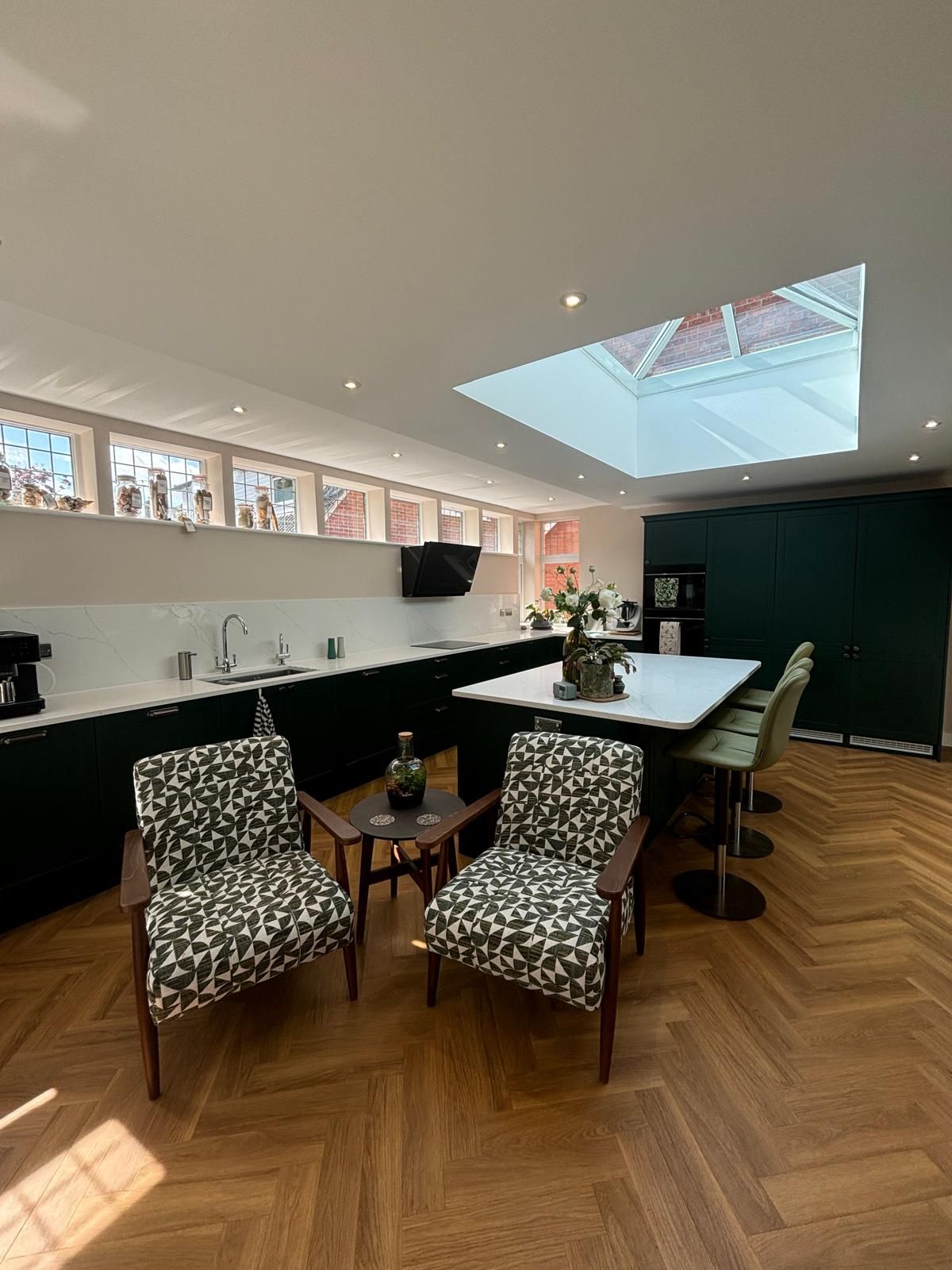 A modern kitchen with dark cabinets, a white island with bar stools, two patterned armchairs, and a large roof lantern.