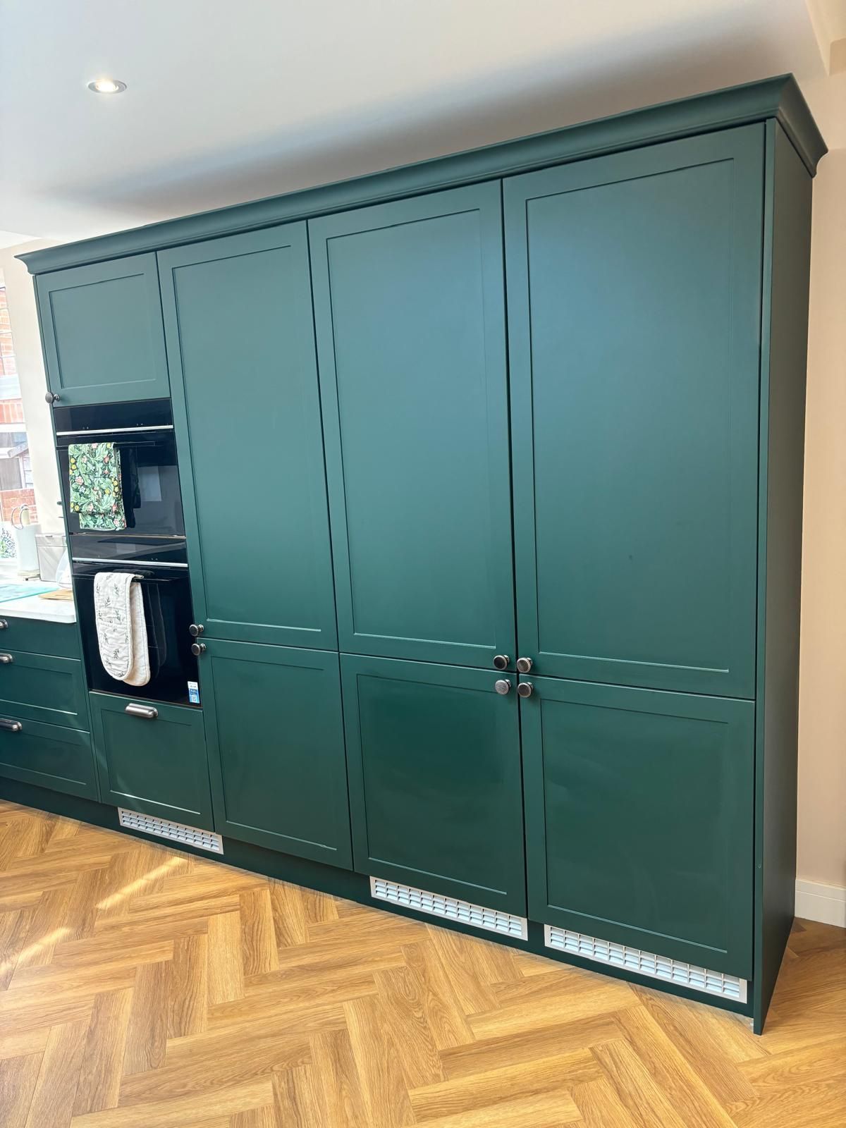 A row of tall, dark green kitchen cabinets with integrated appliances and silver knobs on a light herringbone wood floor.