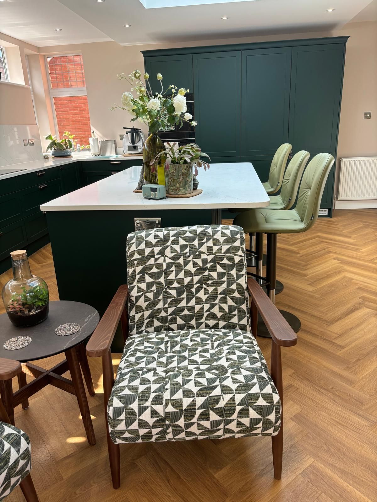 A patterned armchair sits in front of a kitchen island with green cabinets, a white countertop, and light green stools.