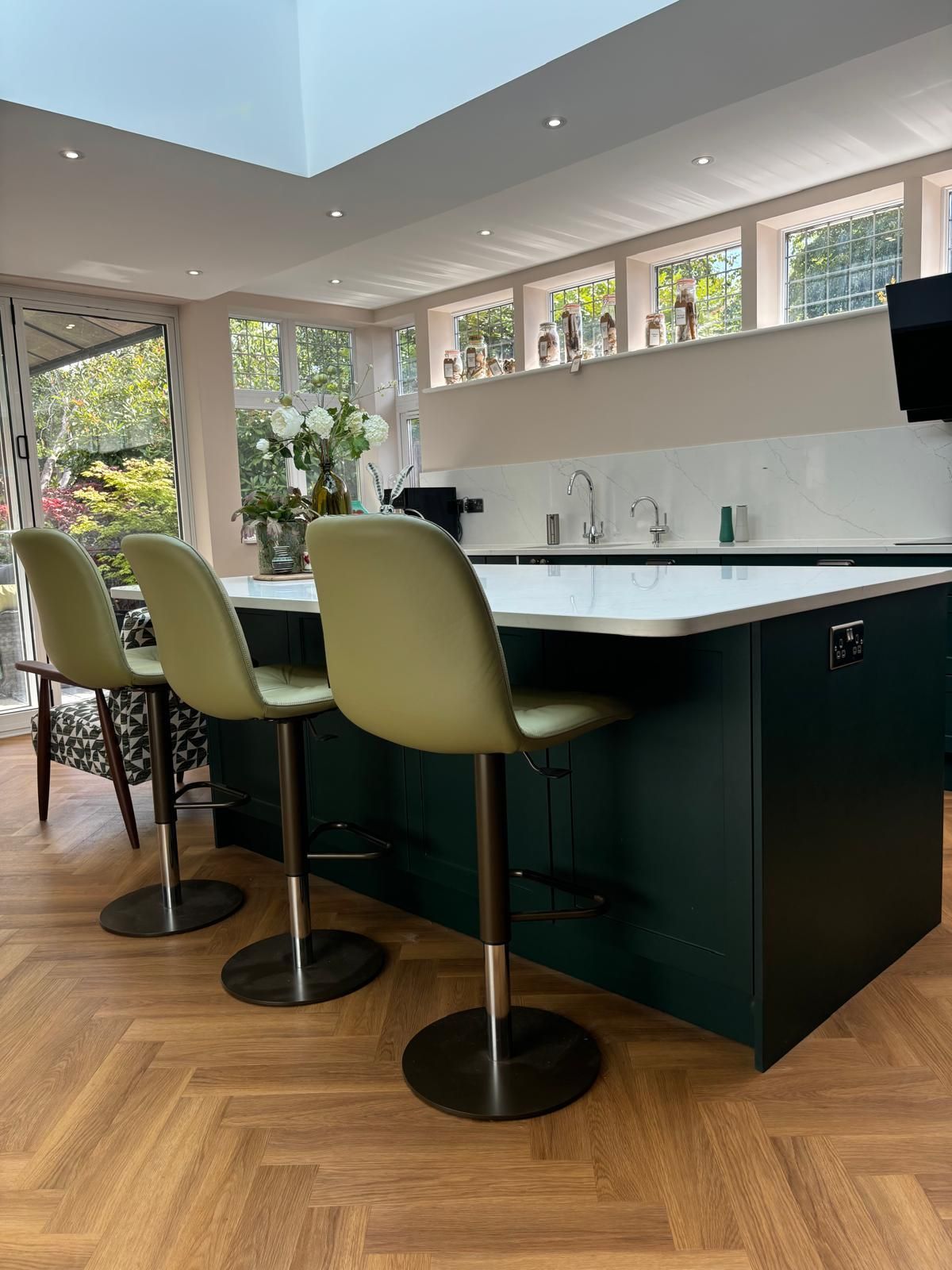 A kitchen island with three sage green bar stools on a herringbone wood floor in a light-filled kitchen.