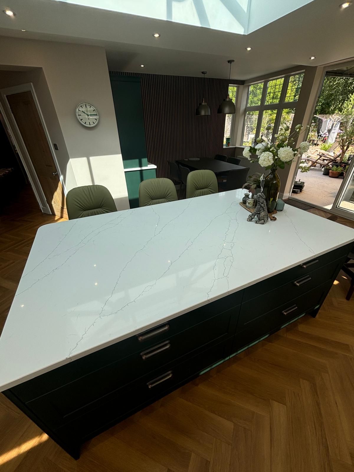 A large white kitchen island with marble-style veining and dark green cabinets, set on herringbone wood flooring.