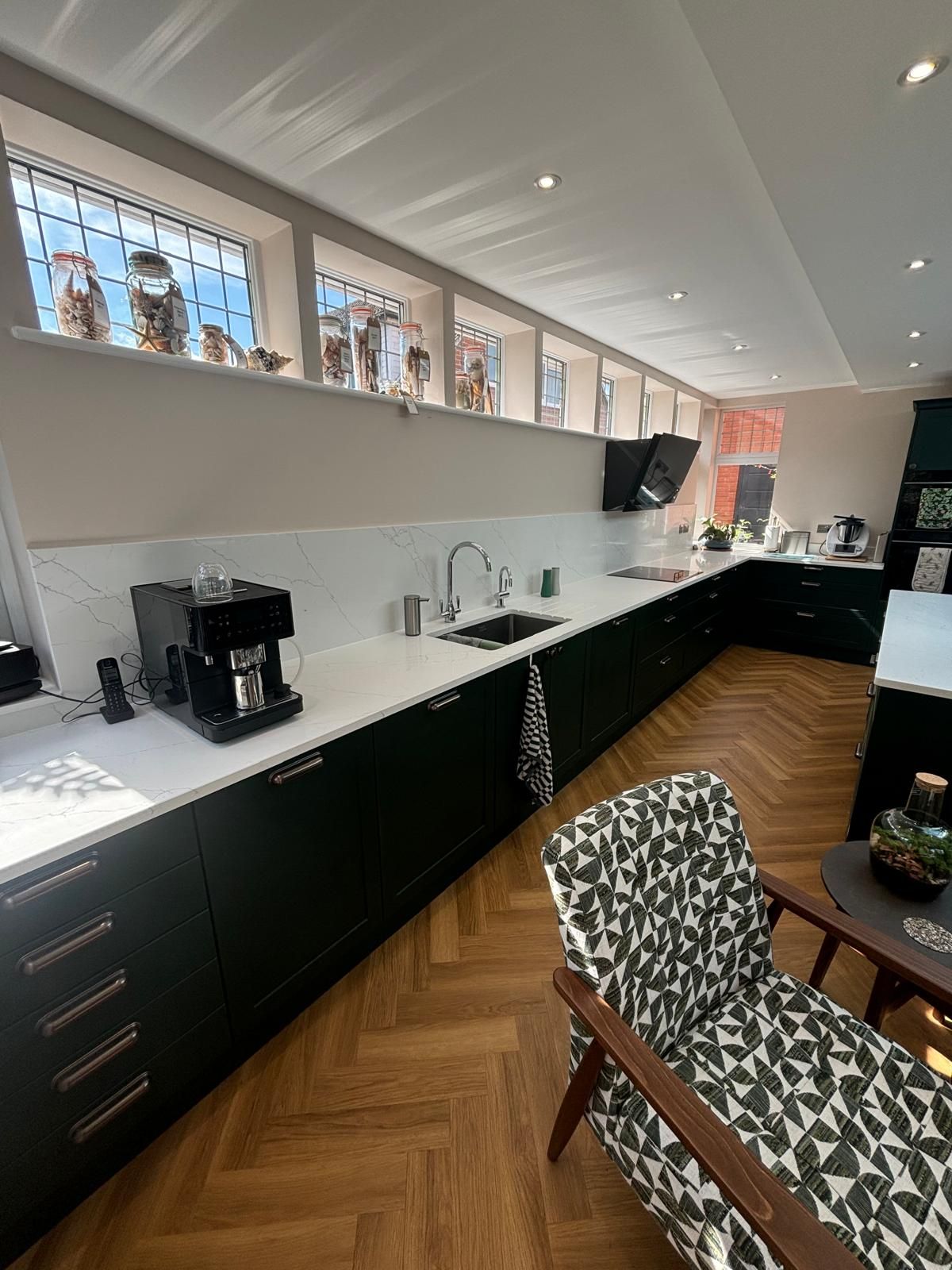 A modern kitchen with dark cabinets, white countertops, herringbone wood floors, and a row of high windows.