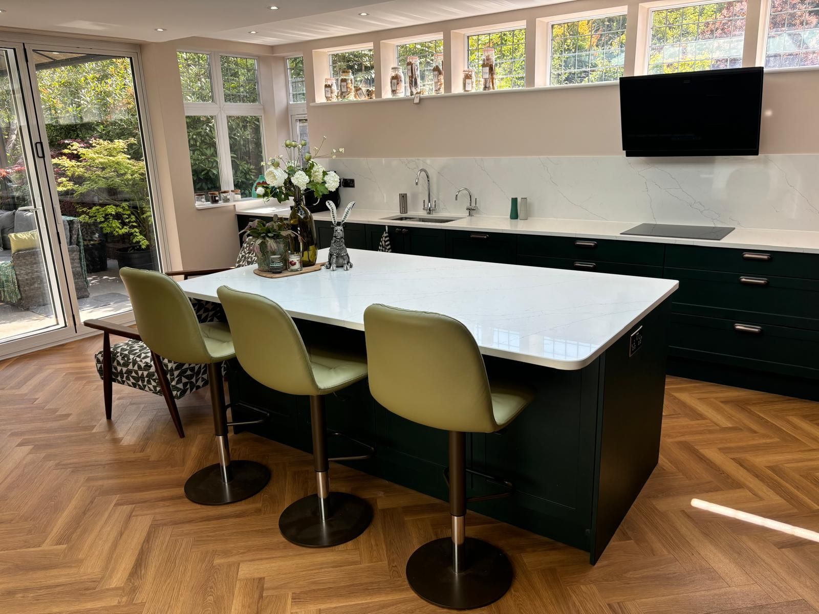 A modern kitchen with a large white island, green chairs, dark green cabinets, and herringbone wood floors.