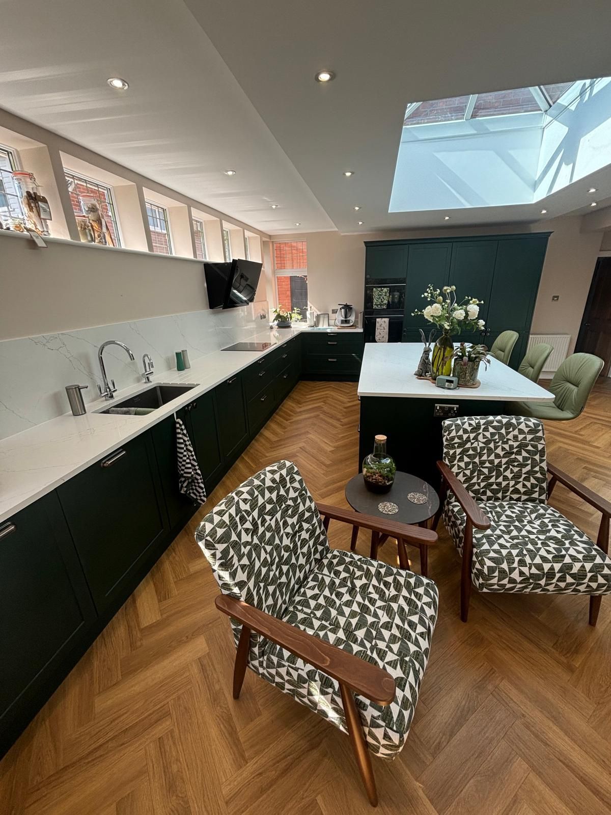 A modern kitchen with dark green cabinets, a white island, two patterned armchairs, and a skylight over hardwood floors.