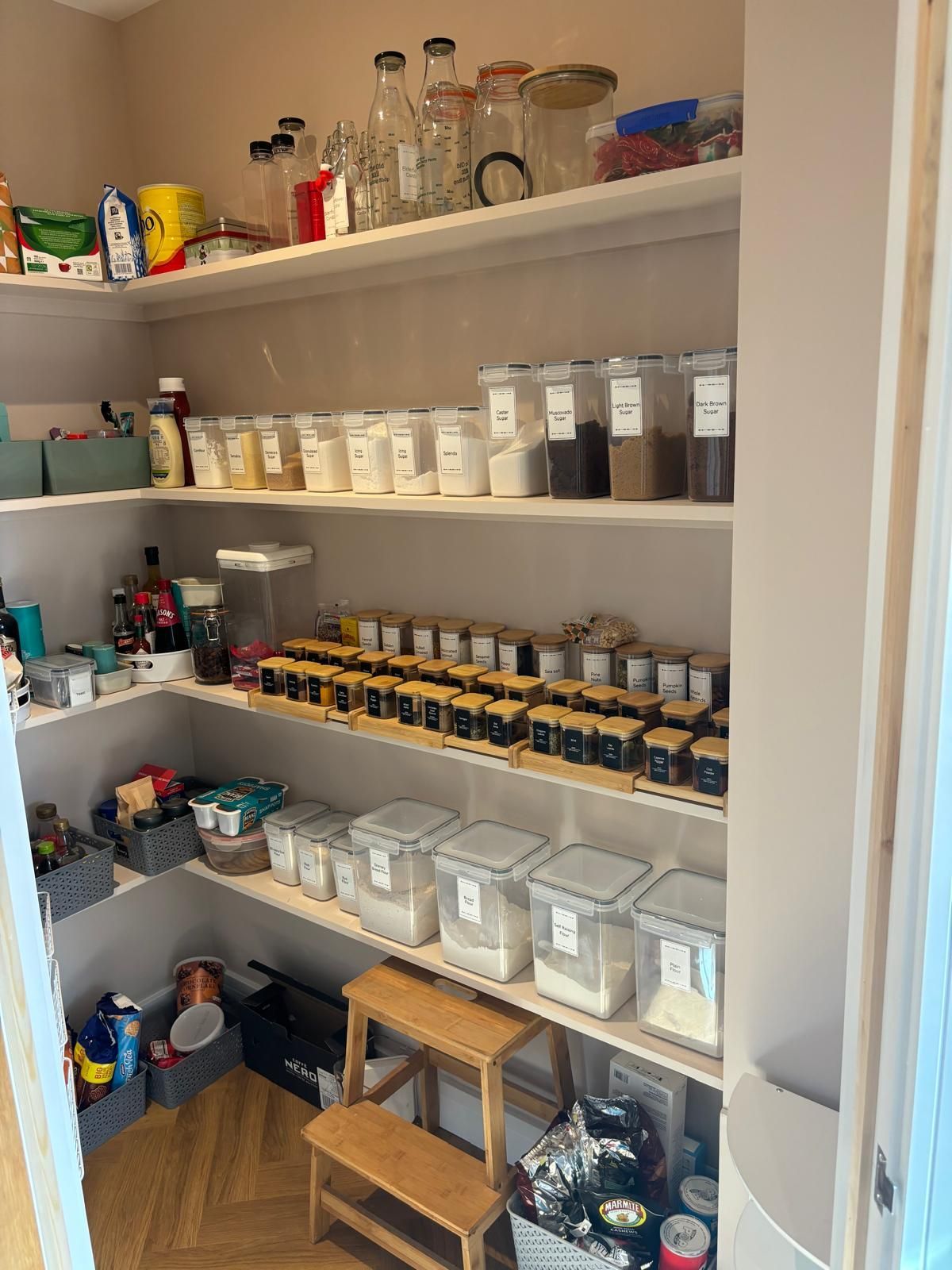 Organized pantry with shelves filled with clear storage containers, glass jars, spices, and a wooden step stool.