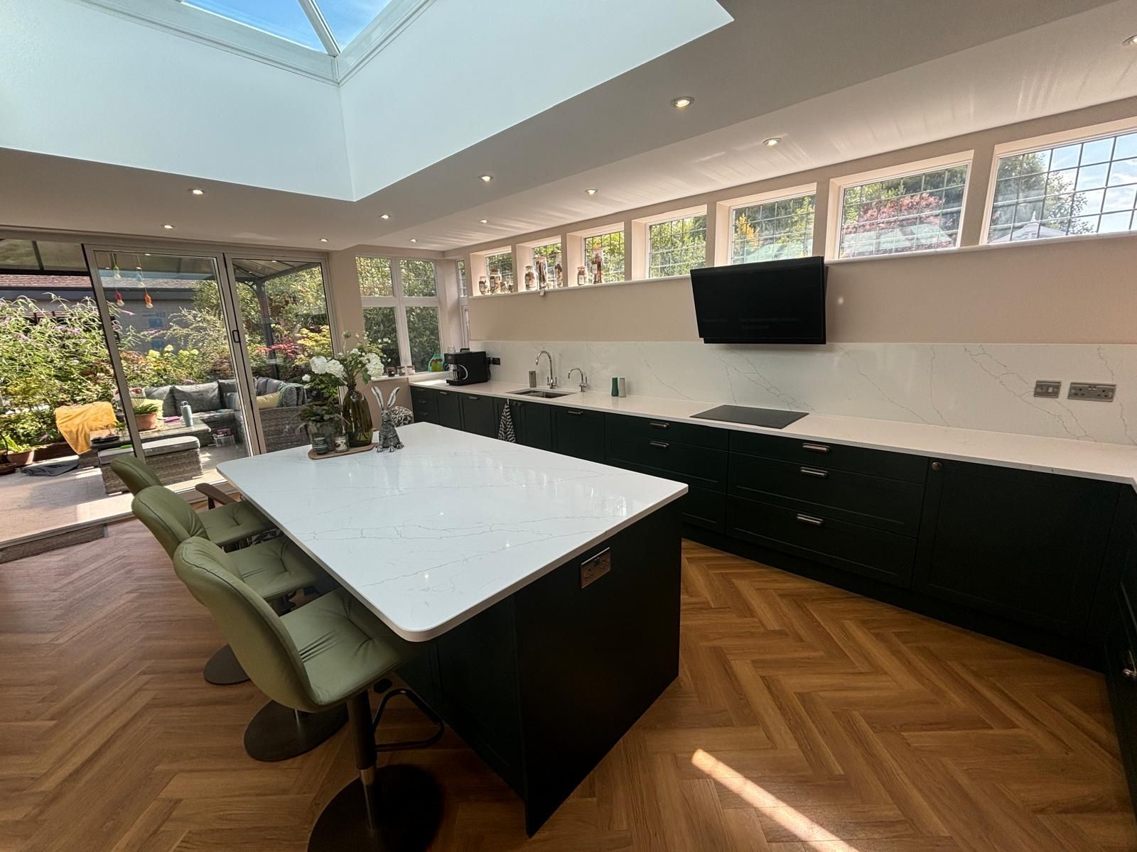 A modern kitchen with a white island, sage green bar stools, black cabinets, wood floors, and a glass skylight ceiling.