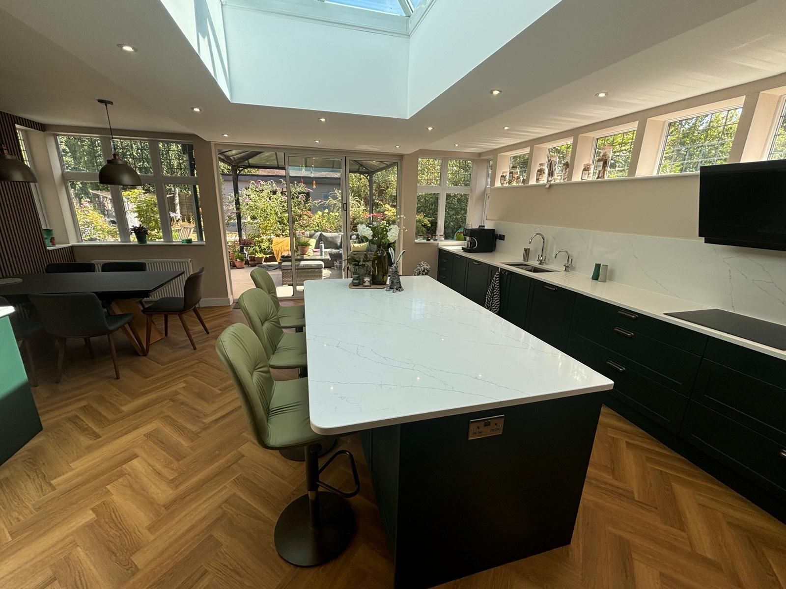 A modern kitchen with dark green cabinets, a large white kitchen island, three olive chairs, and light herringbone floors.