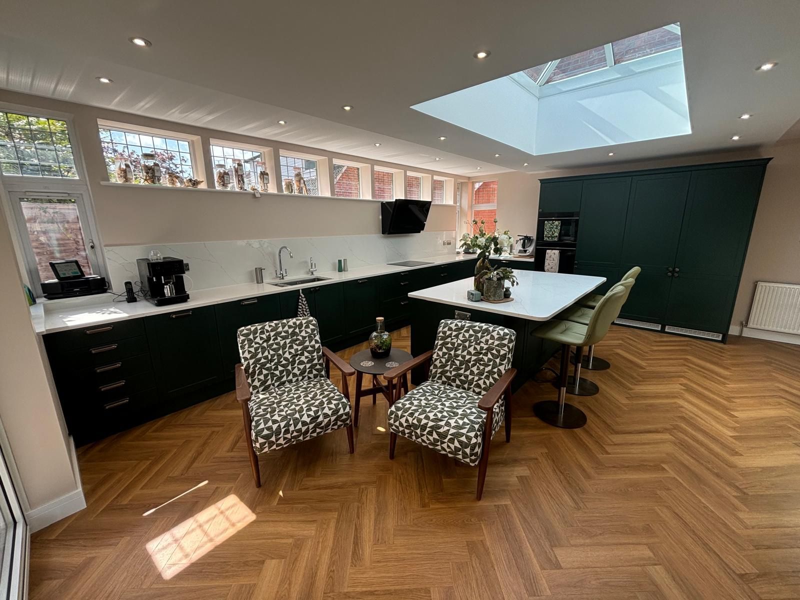 Modern kitchen with green cabinets, a kitchen island, patterned chairs, and a herringbone wooden floor under a skylight.