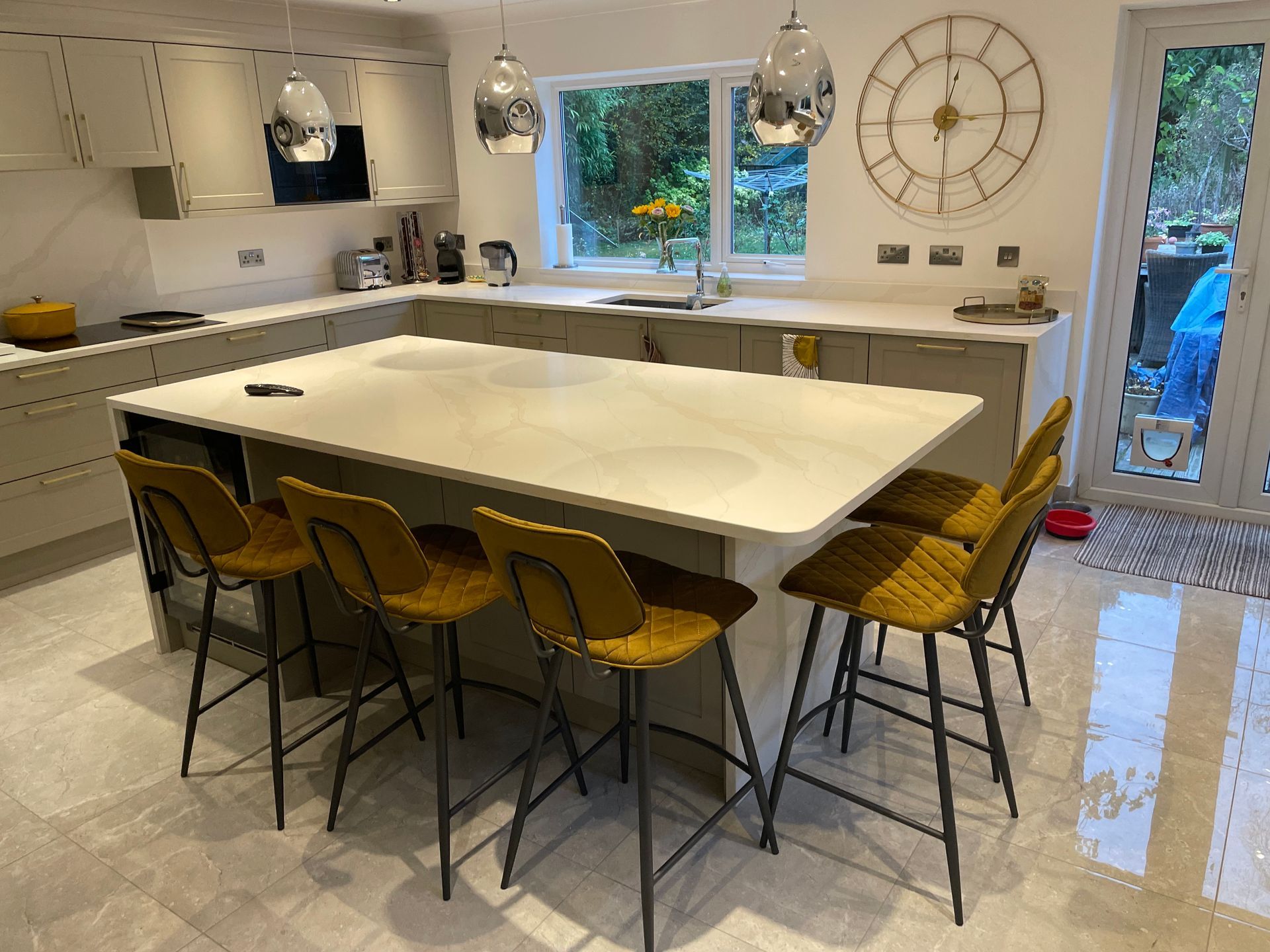 Modern kitchen with a large white island, four yellow bar stools, neutral cabinets, and a large wall clock.