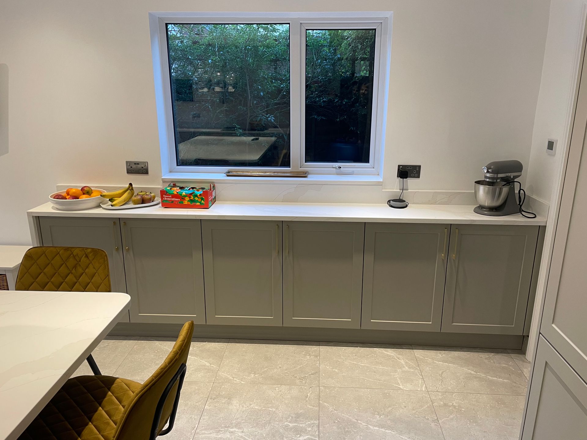 A kitchen with long grey cabinets under a window, featuring a stand mixer, fruit bowls, and mustard-colored dining chairs.
