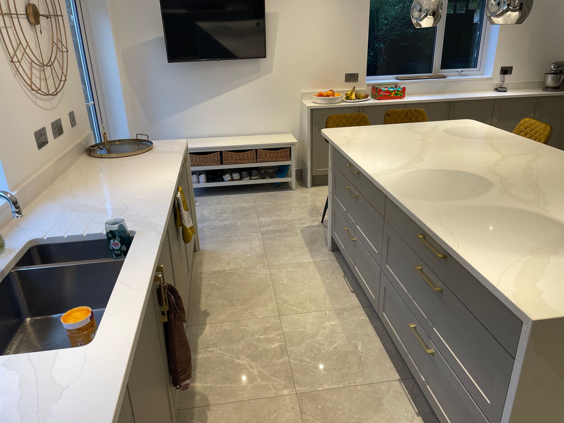 A modern kitchen with grey cabinetry, a large white stone island, tiled floors, and a wall-mounted television.