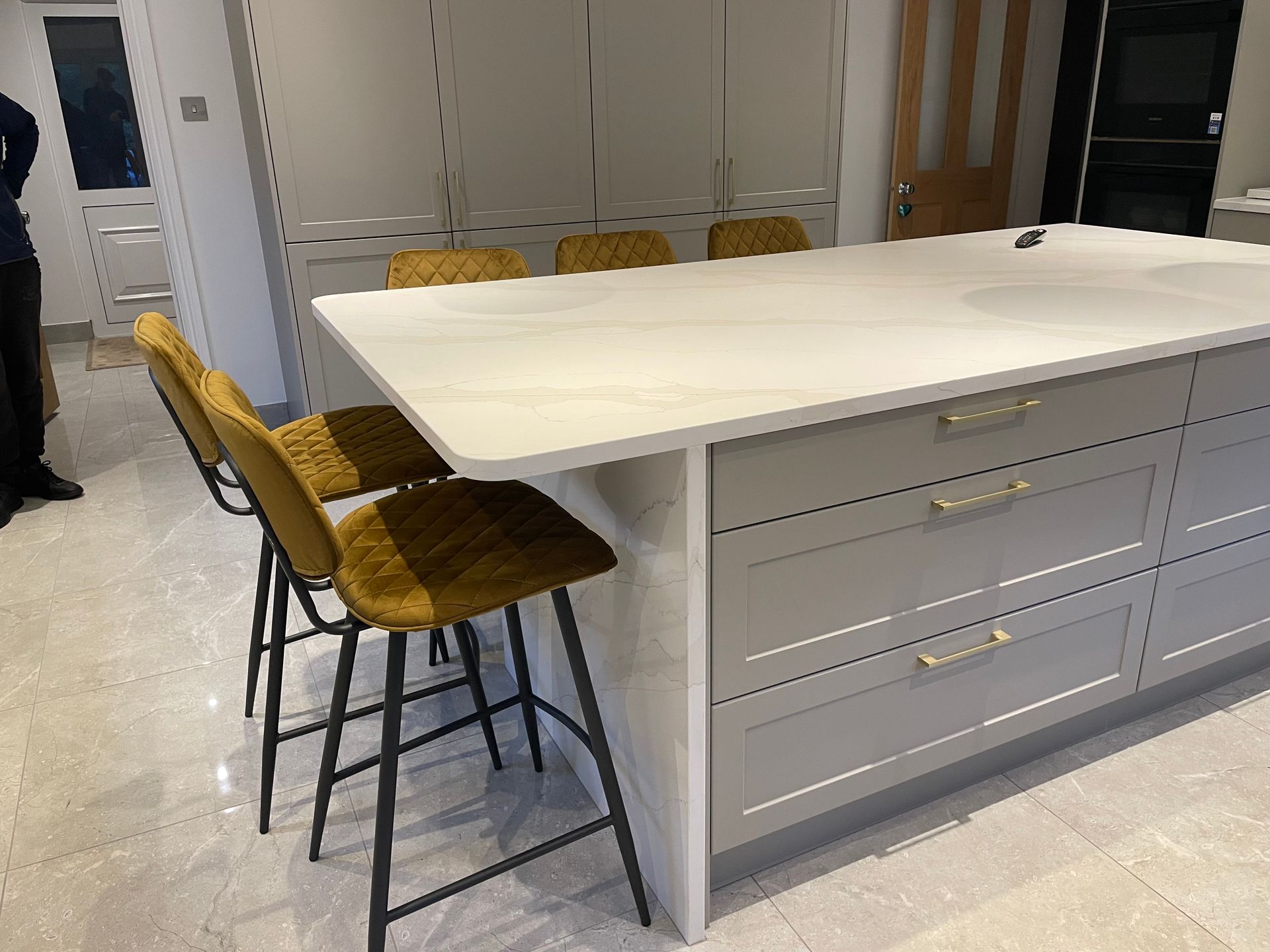 A kitchen island with a white countertop and grey cabinets, featuring three yellow velvet stools on a marble floor.