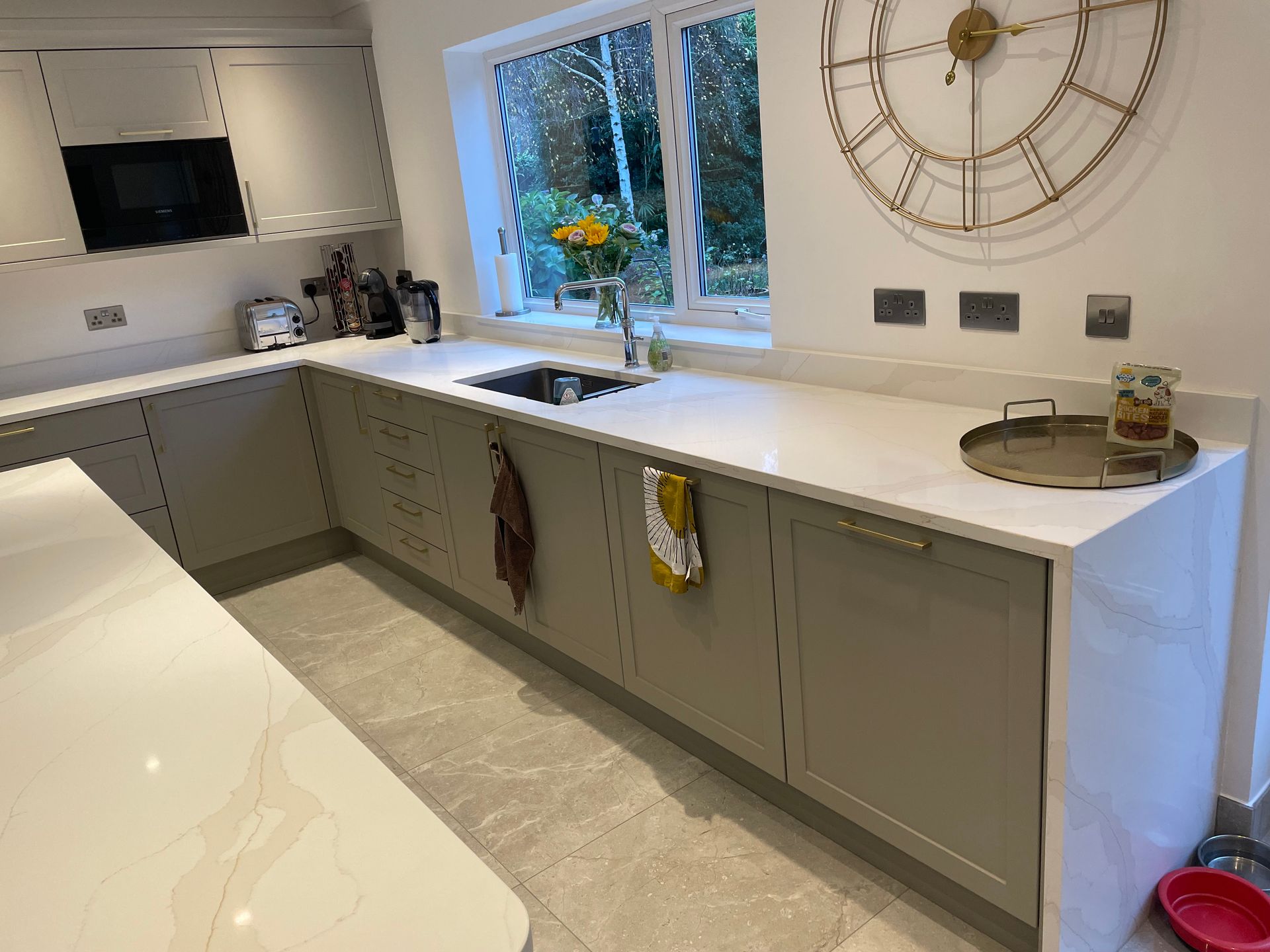 A modern kitchen featuring grey cabinets, white countertops, a sink under a window, and a large decorative wall clock.