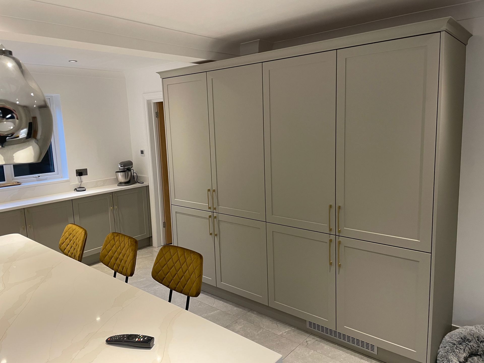 A tall, beige, shaker-style cabinetry unit stands next to a kitchen island with three mustard-yellow bar stools.