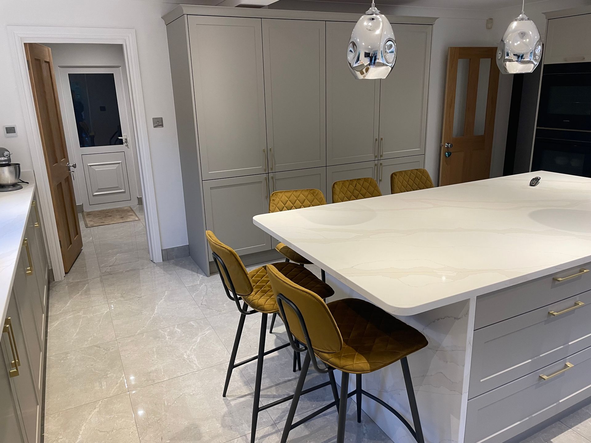A modern kitchen with light grey cabinets, a white island, and three gold bar stools on tiled floors.