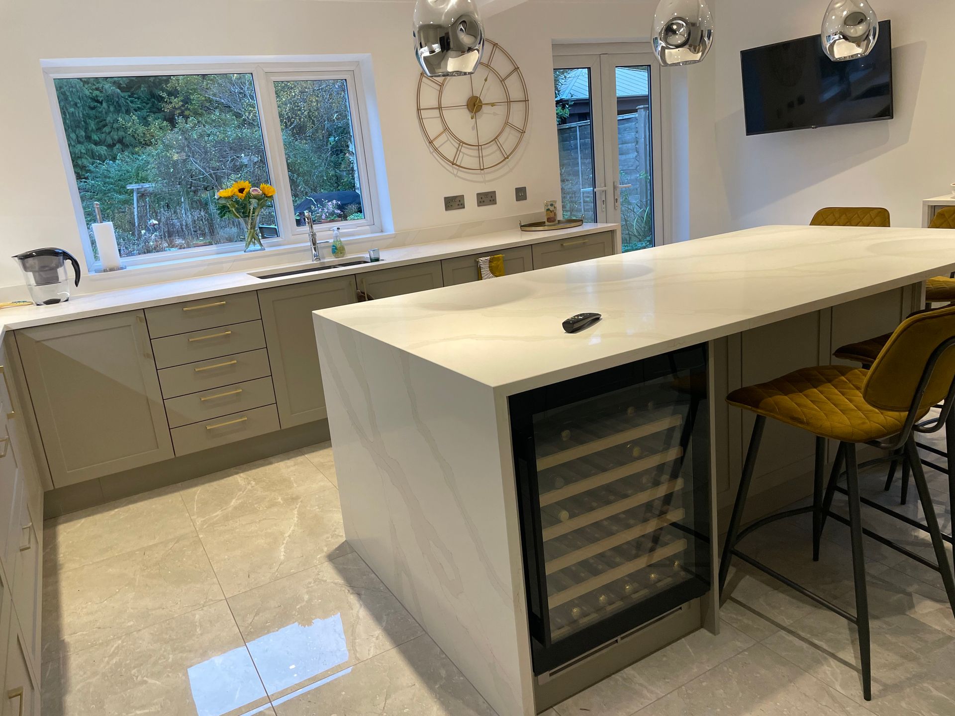 A modern kitchen featuring light grey cabinets, a large white island with a wine cooler, and mustard-colored bar stools.