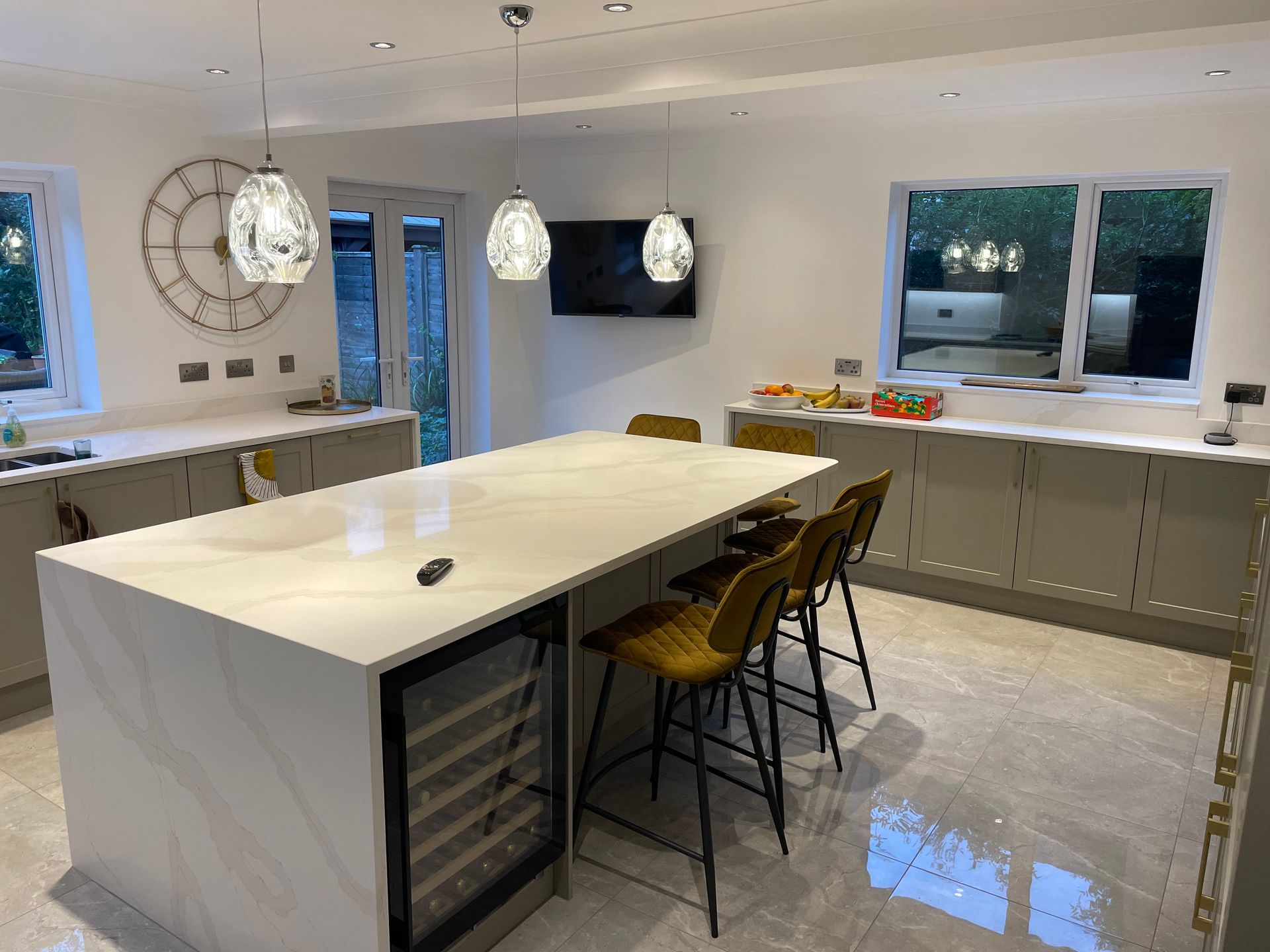 Modern kitchen featuring a large white quartz island with three mustard-colored stools, light gray cabinetry, and lighting.