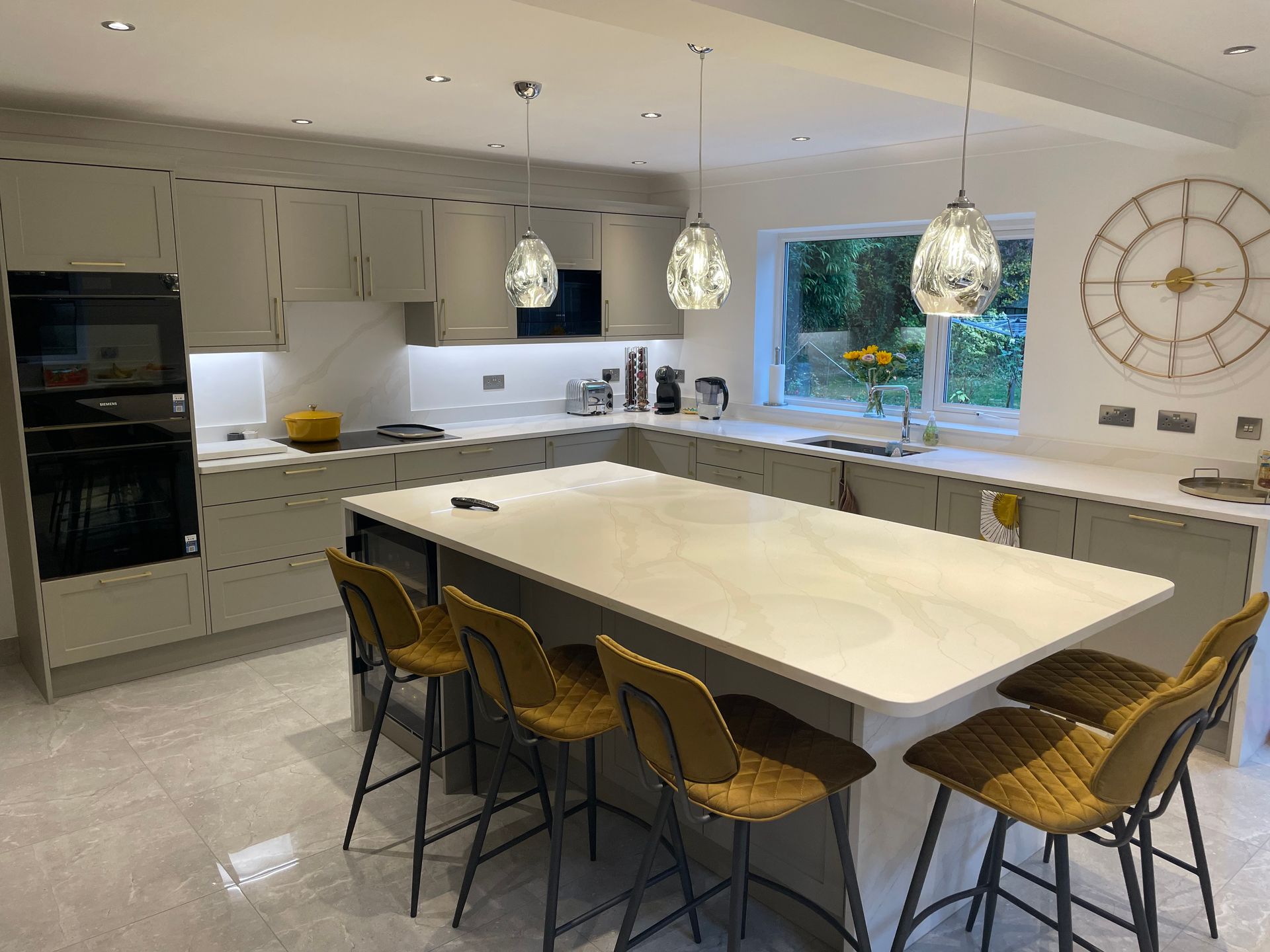 A modern kitchen with light grey cabinets, a large central island with four mustard-yellow bar stools, and a wall clock.
