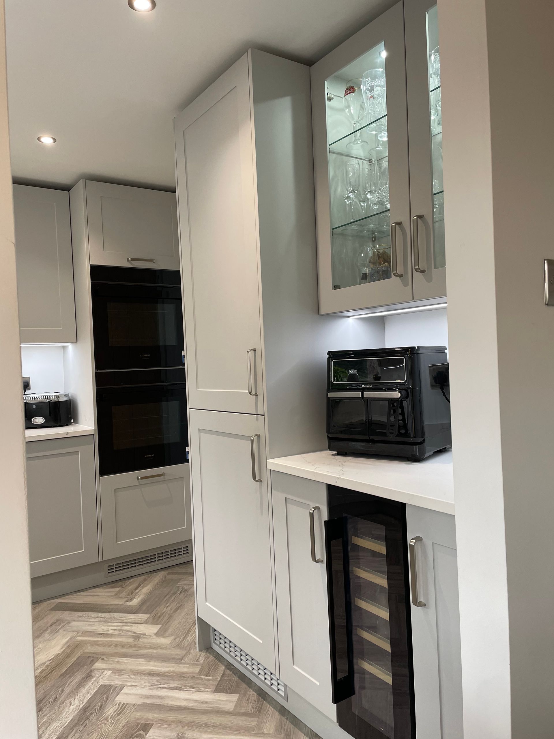 Narrow modern kitchen with white cabinets, built-in oven, and dark appliances along a light wood floor