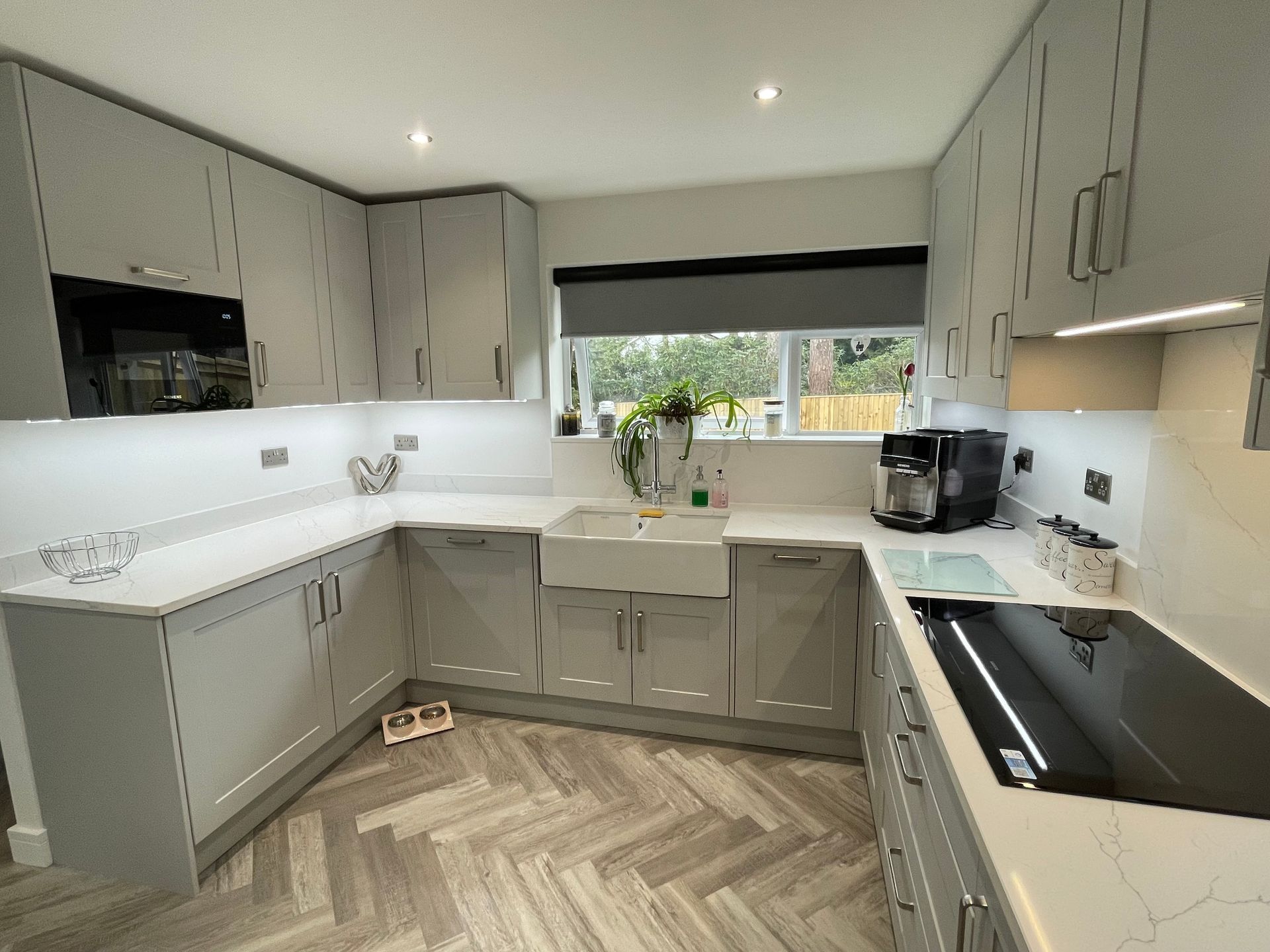 Bright modern kitchen with gray cabinets, white counters, herringbone floor, and a window over the sink.