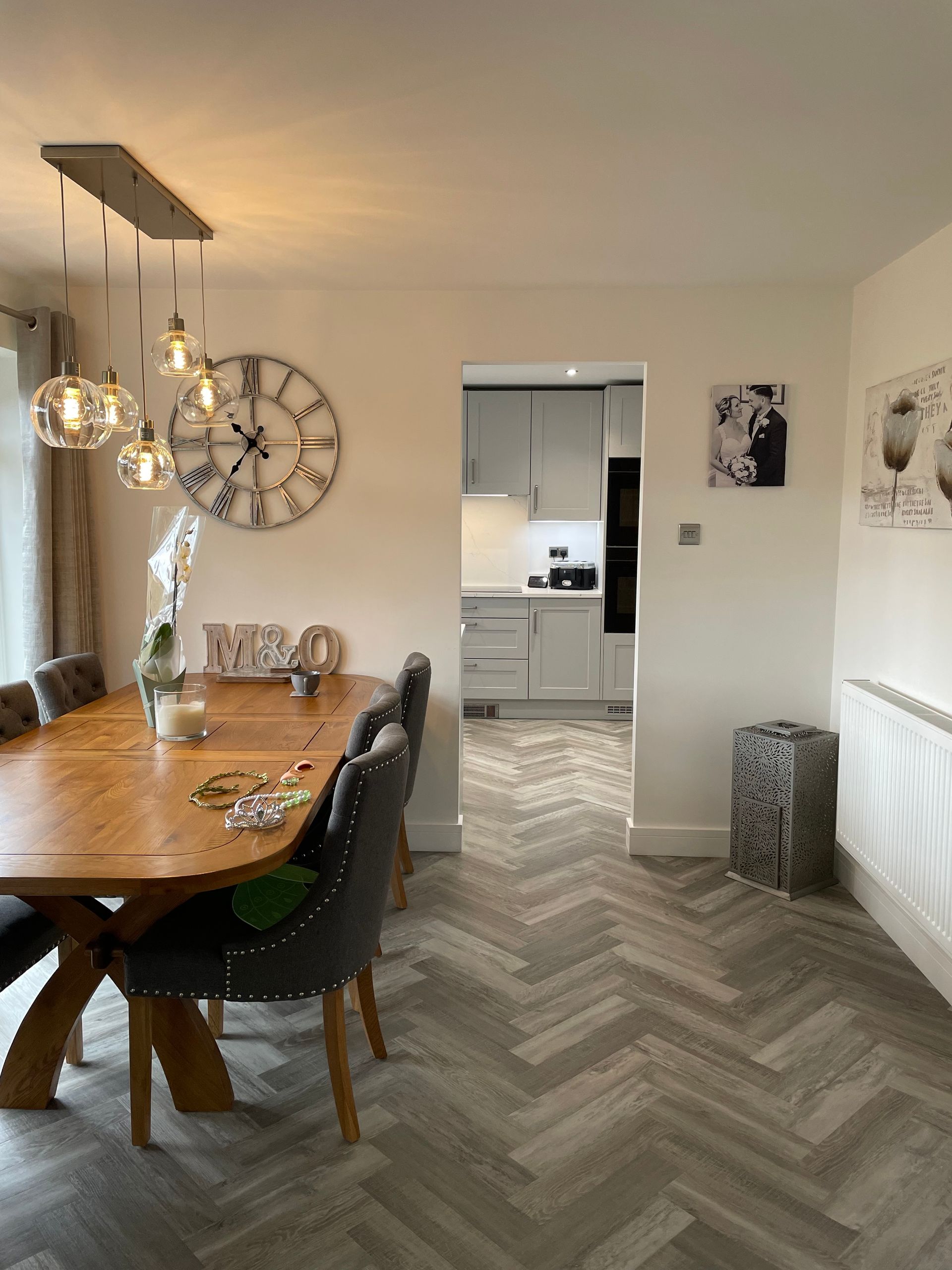 Modern dining room with wooden table, hanging lights, wall clock, and herringbone-patterned floor.