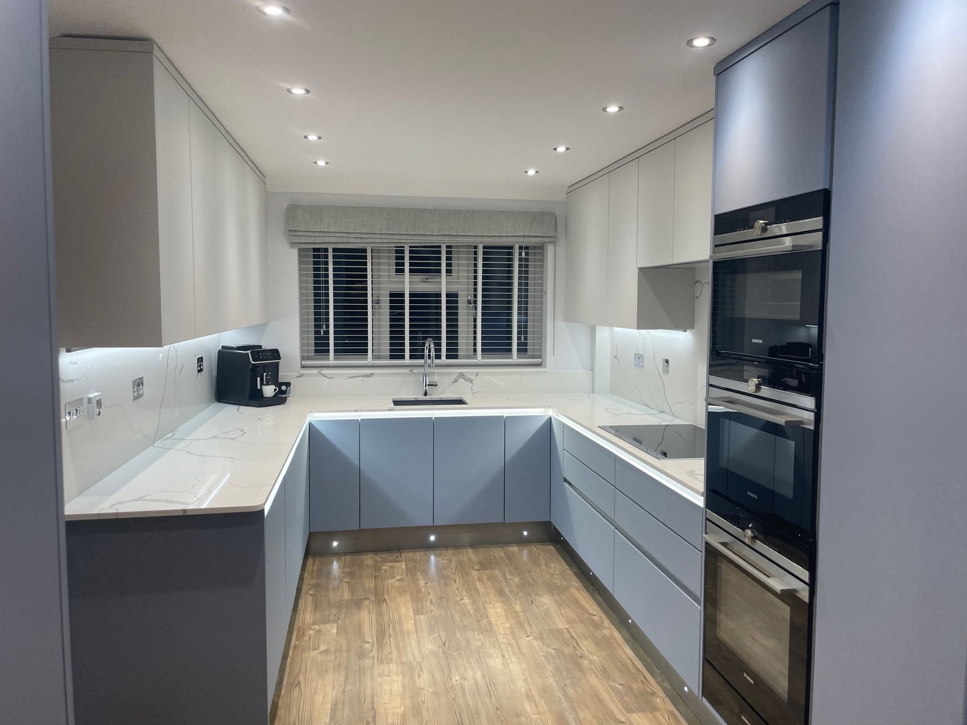 A modern U-shaped kitchen with light gray cabinets, white countertops, stainless steel wall ovens, and wood-look flooring.